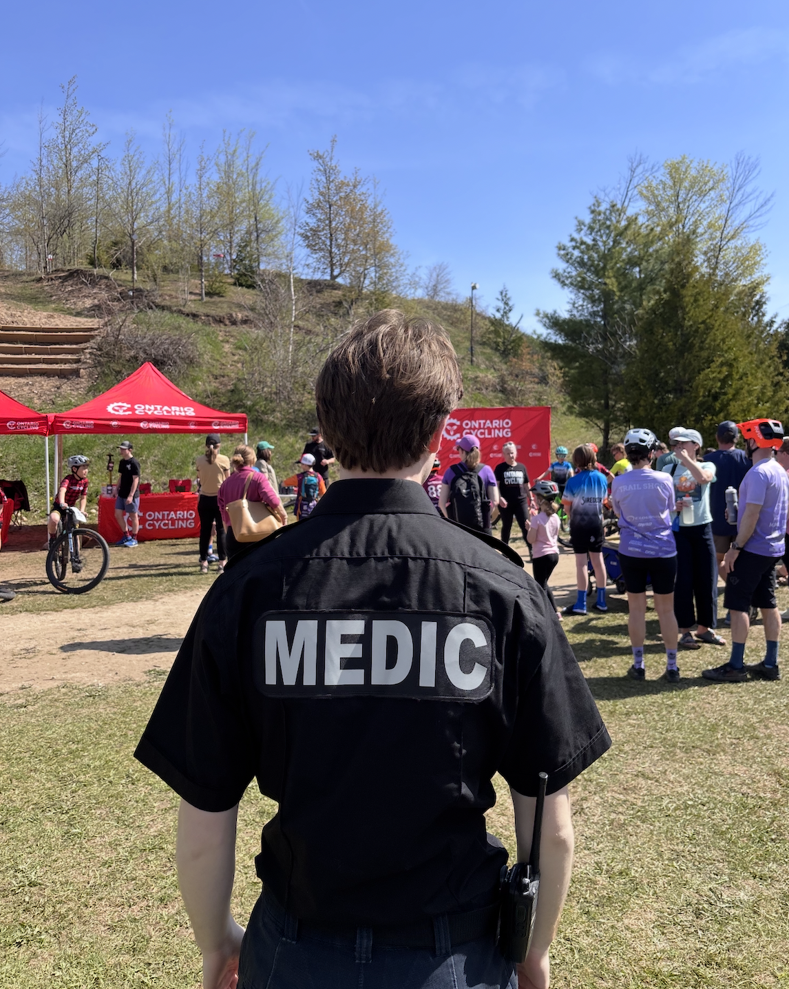 A medic stands in the foreground at a cycling event, with a crowd gathered around tents and a stage in the background, under a partly cloudy sky with trees on a grassy field.