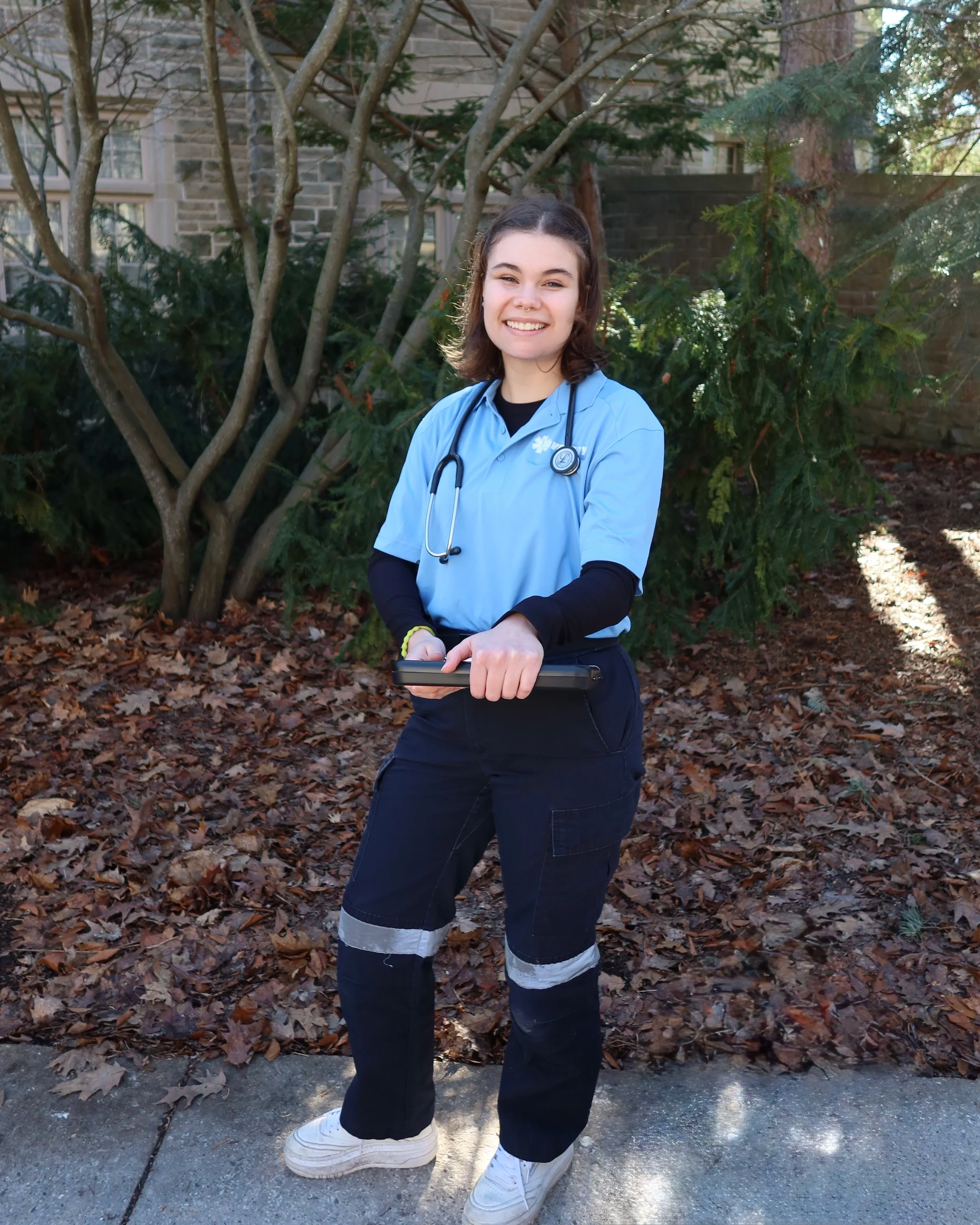 A young female nurse in medical scrubs with a stethoscope around her neck holding a tablet, standing outdoors in front of trees and a house.