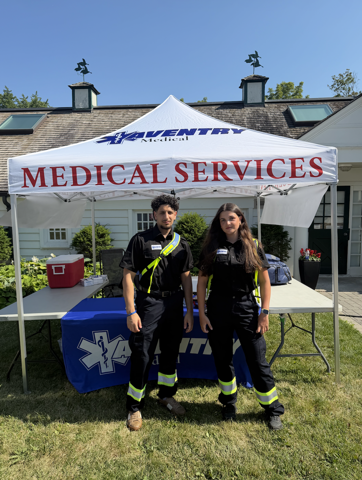 Two Aventry emergency medical technicians standing under a tent with a sign that reads Medical Services. They are outdoors in front of a house with a garden and sunny sky.