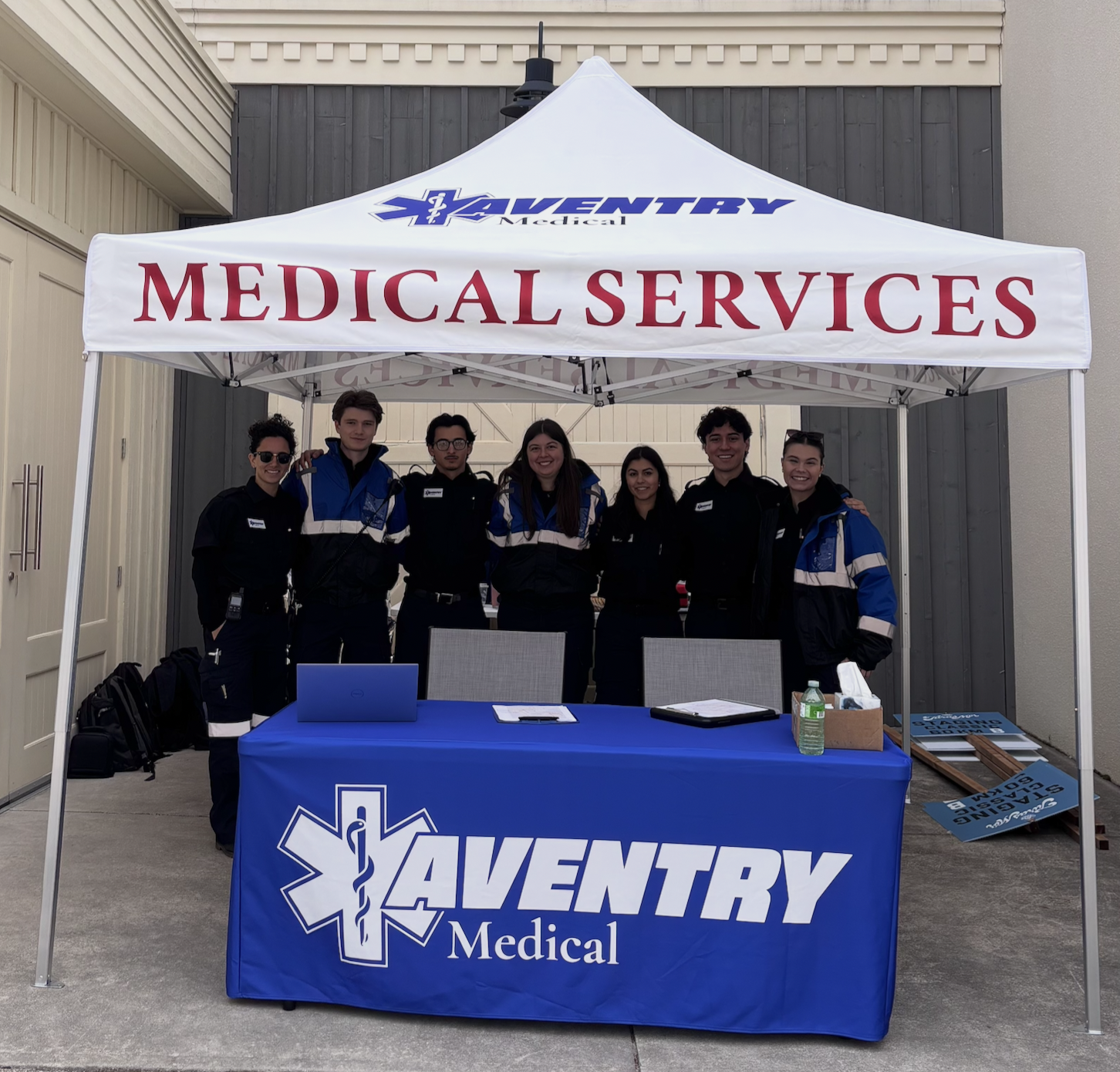 Group of seven Aventry Medical medics standing behind a table at a community event, under a white tent with 'Medical Services' and 'AVENTRY Medical' signage.