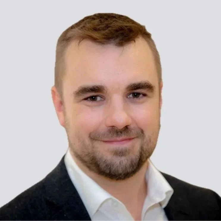 Portrait of Dr. Frank Myslik with short brown hair, a beard, and mustache, wearing a white shirt and dark blazer, smiling softly against a blurred background.