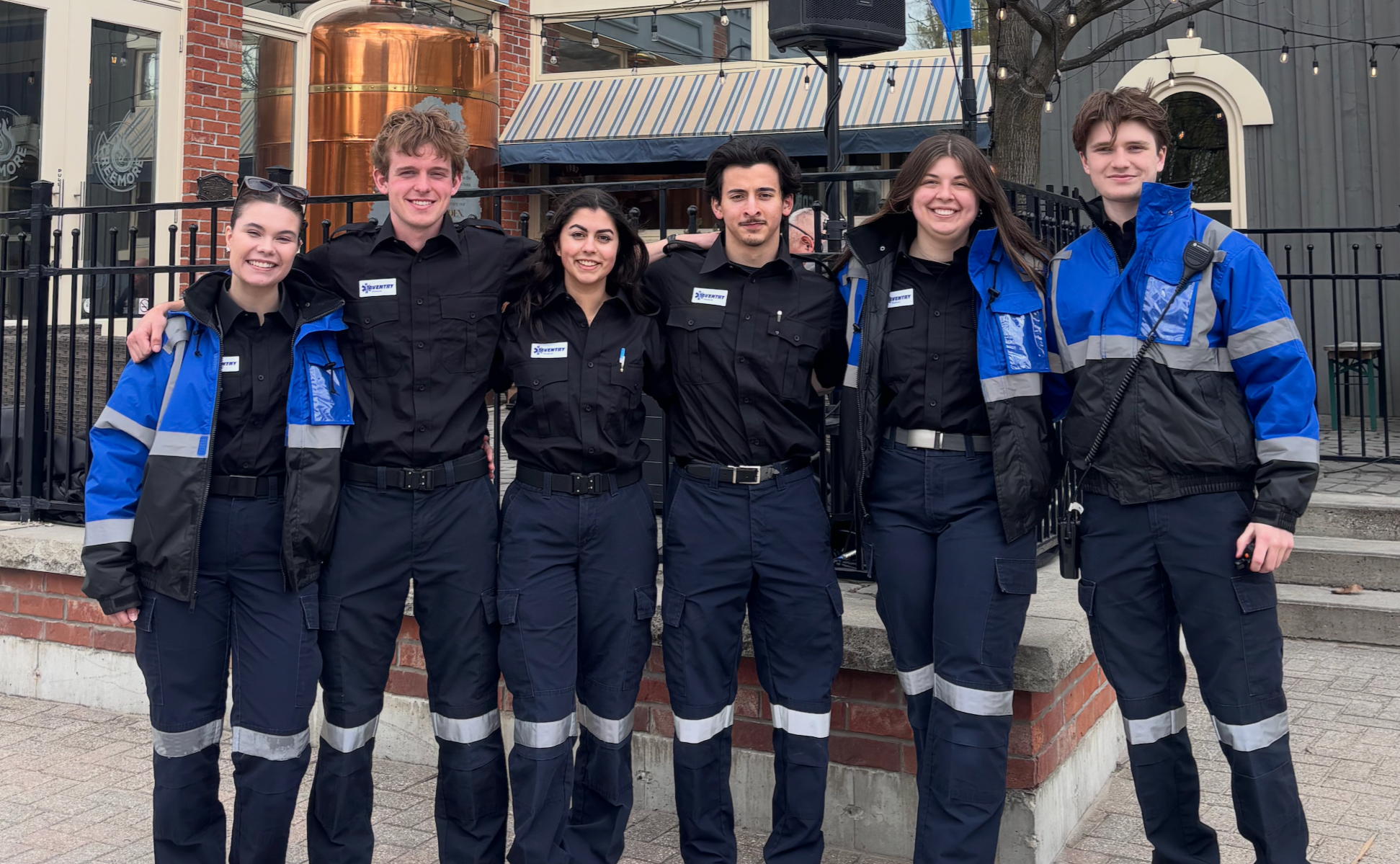 Group of six young people in uniform standing outdoors in front of a brick and black building with string lights above, posing for a photo.