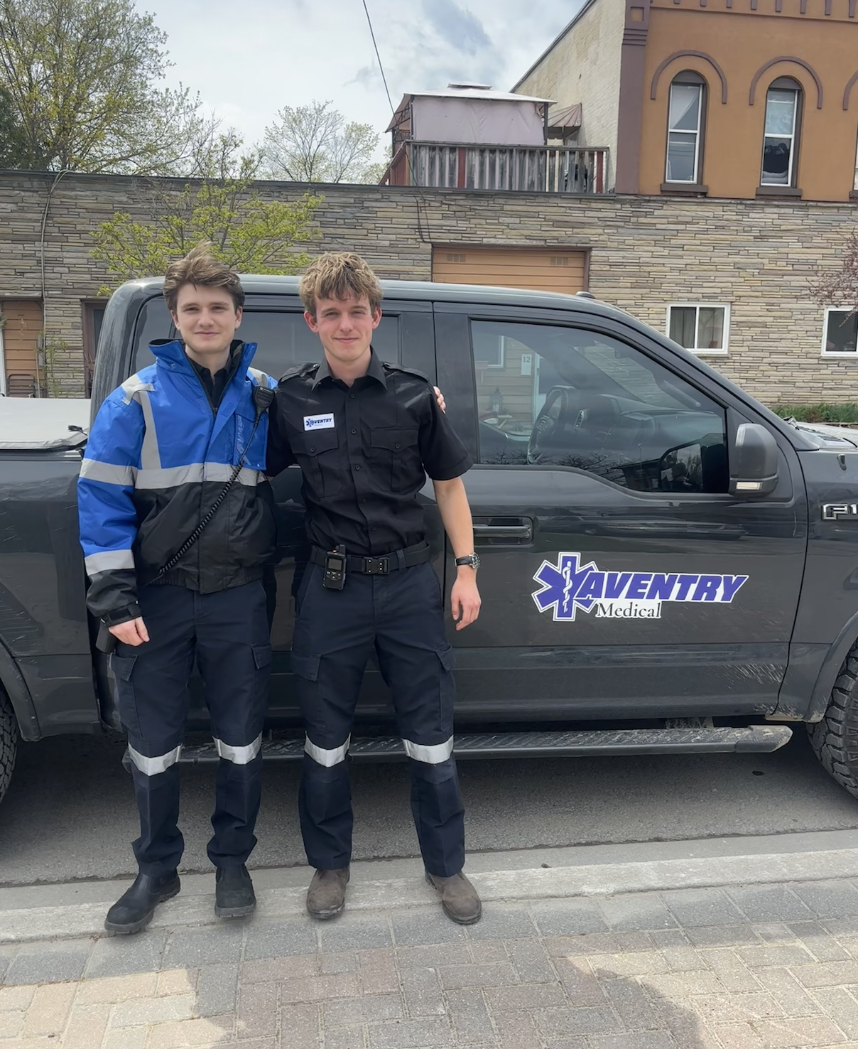 Two young men in Aventry emergency medical uniforms standing in front of a black vehicle with 'Aventry Medical' logo. They are smiling and arm in arm, outdoors in a residential area.
