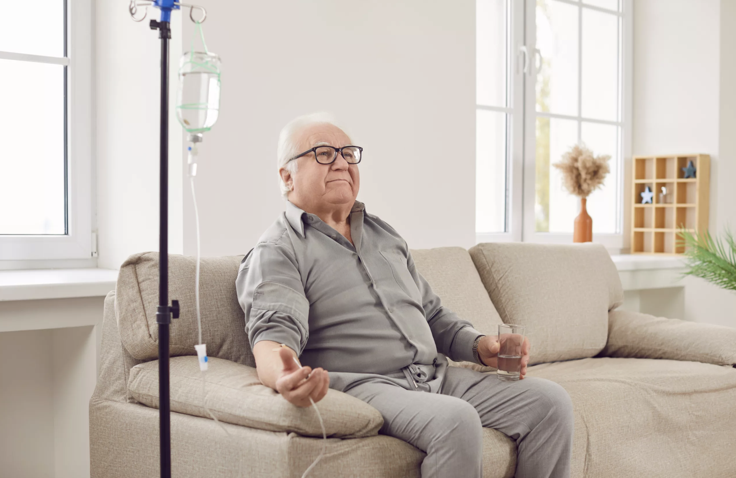 An elderly man sitting on a beige couch in a bright room with a window, holding a glass of water in one hand, with an IV stand beside him.