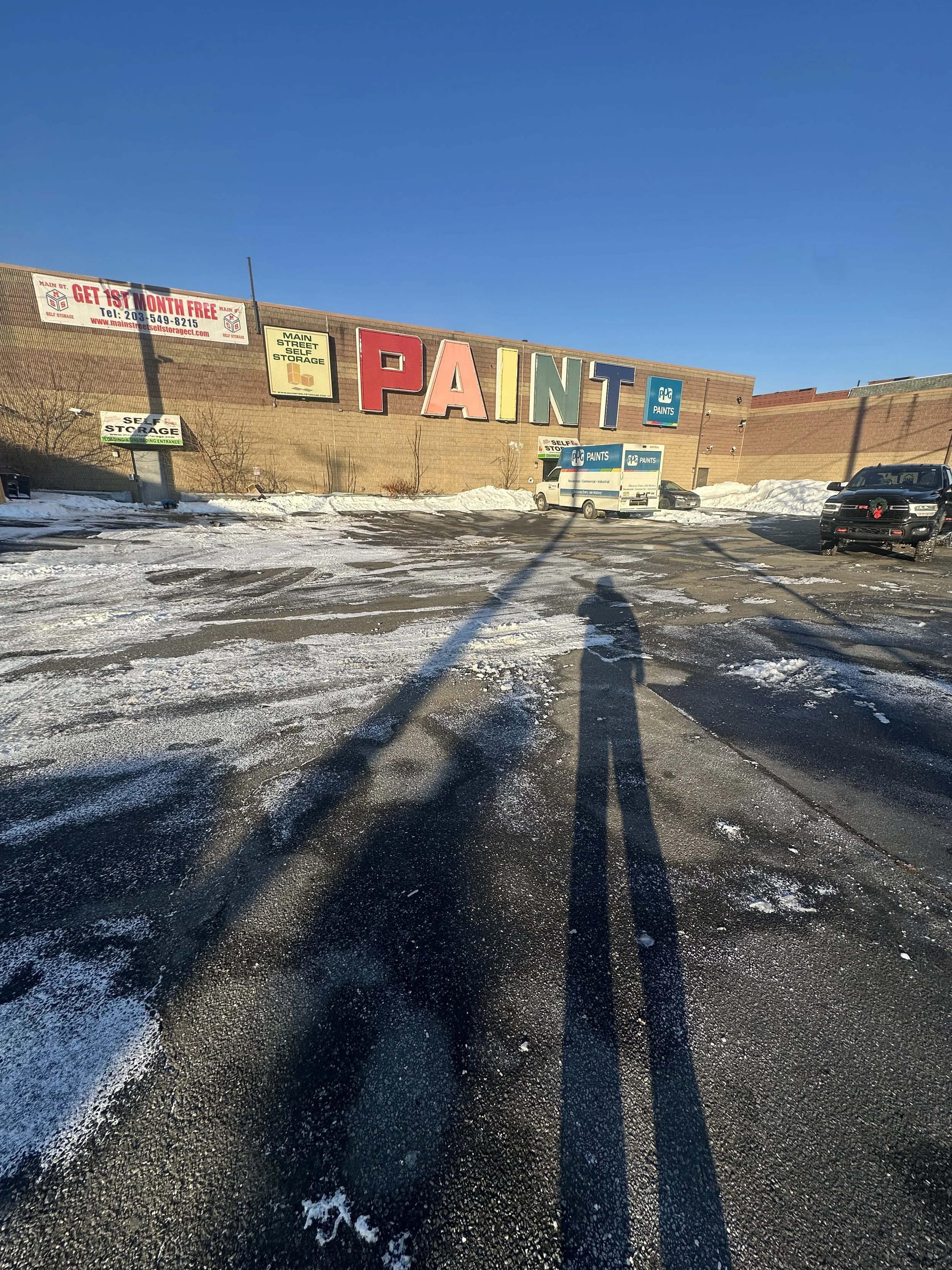 A parking lot with snow and ice, reflecting long shadows of two people taking a photo. In the background, a store with a large sign that spells 'PAINT' and smaller signs advertising storage and paint services.