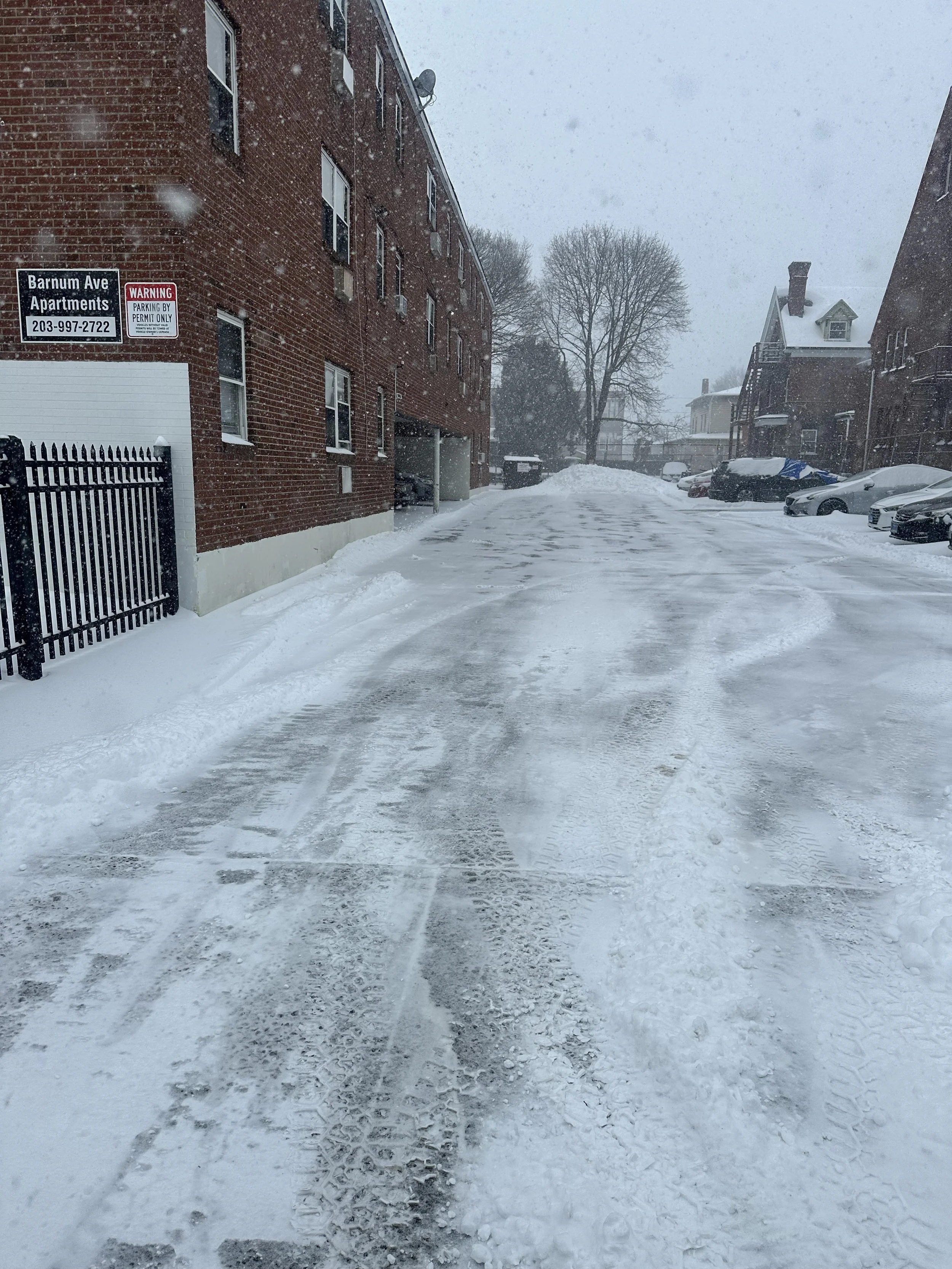 Snow-covered parking lot and street in a residential area with apartment building on the left and houses on the right; snow falling heavily.