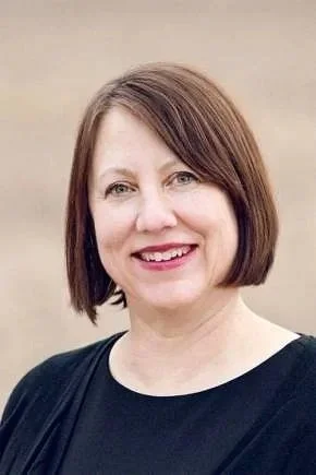 A woman with shoulder-length brown hair smiling, wearing a black top, against a neutral background.