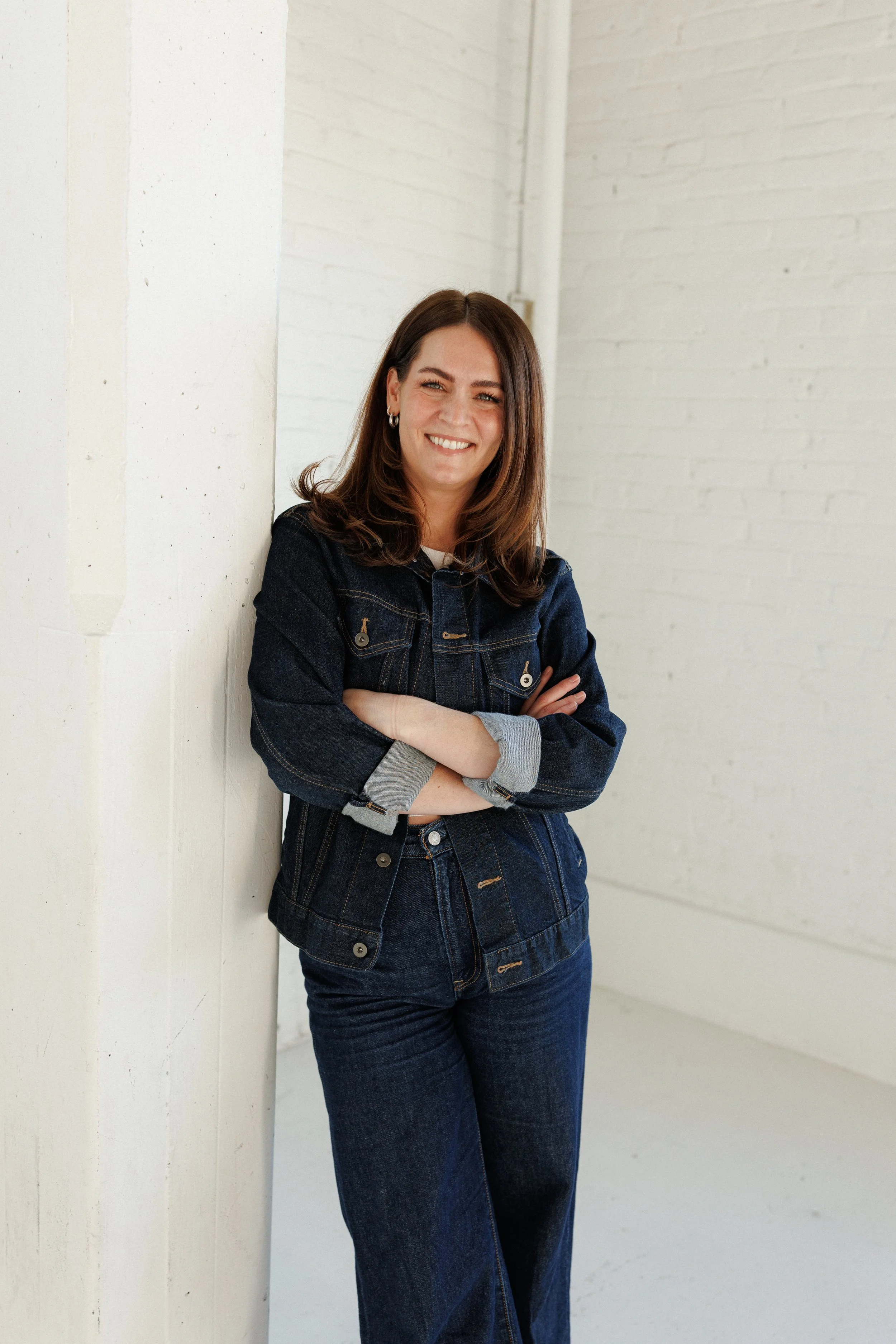 A woman with brown hair smiling, wearing a dark denim jacket and jeans, standing against a white wall with her arms crossed.