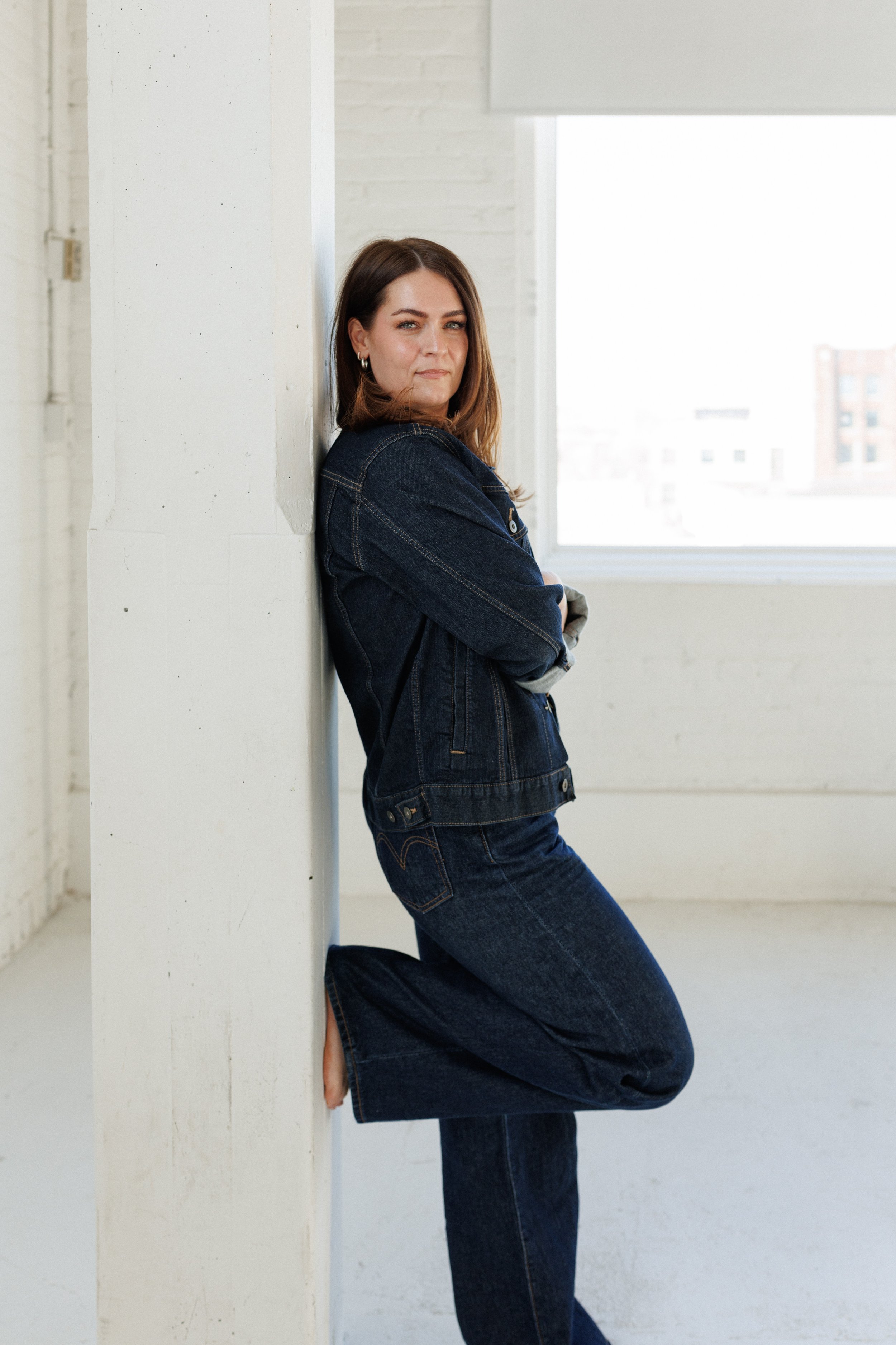 A woman with shoulder-length brown hair, wearing a denim jacket and jeans, leans against a white wall in a bright room with large windows.