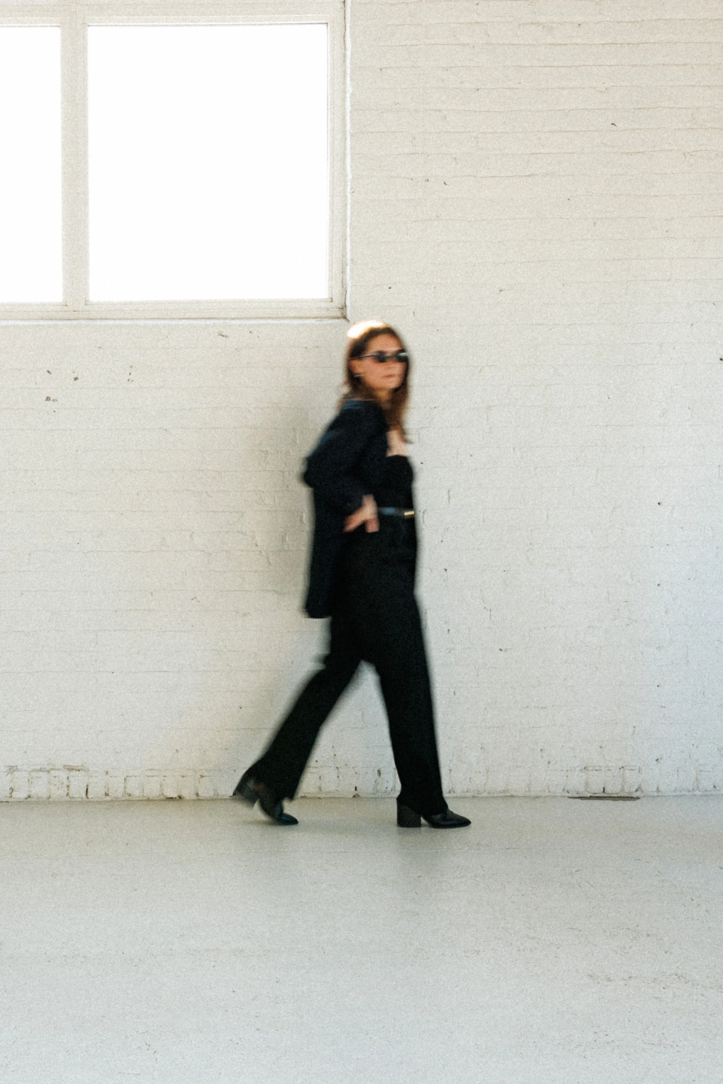 Sara Harrington with Her Money House walking in front of a white brick wall with a large window, wearing black pants, black heels, a black jacket, and sunglasses.