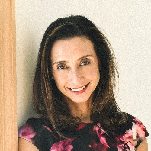 A woman with dark brown hair, smiling, wearing a floral dress, standing indoors near a light-colored wall.