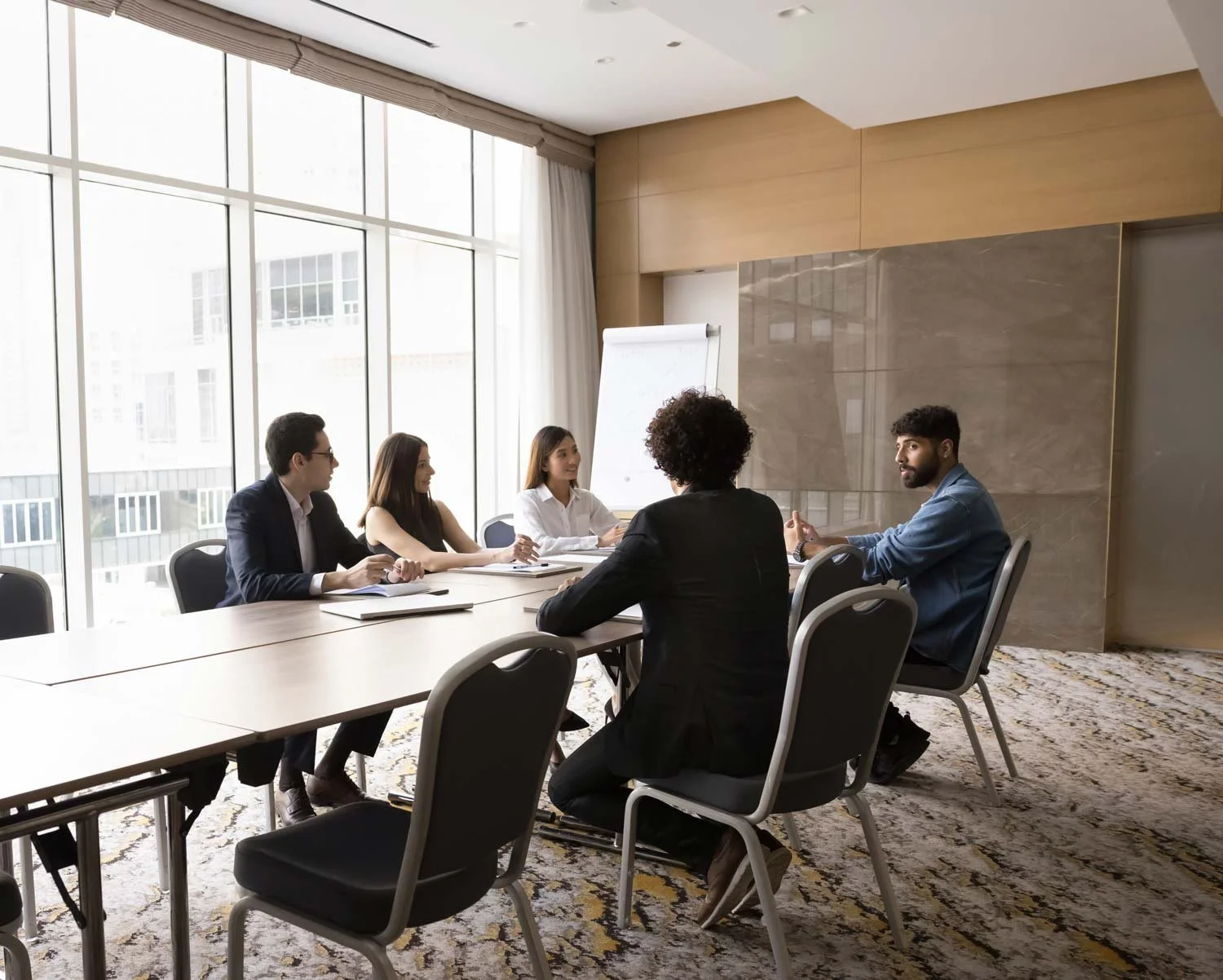 A business meeting with five people seated around a table in a modern, well-lit conference room with large windows and a flip chart.