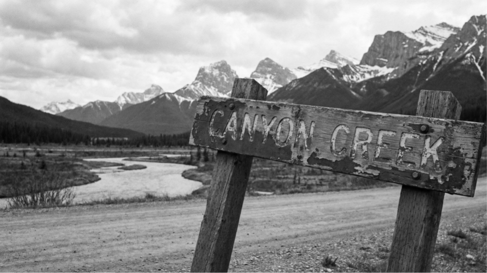 Black and white photograph of a weathered wooden sign that reads 'Canyon Creek,' leaning at an angle in front of a wide river, surrounded by trees, with snow-capped mountains in the background.