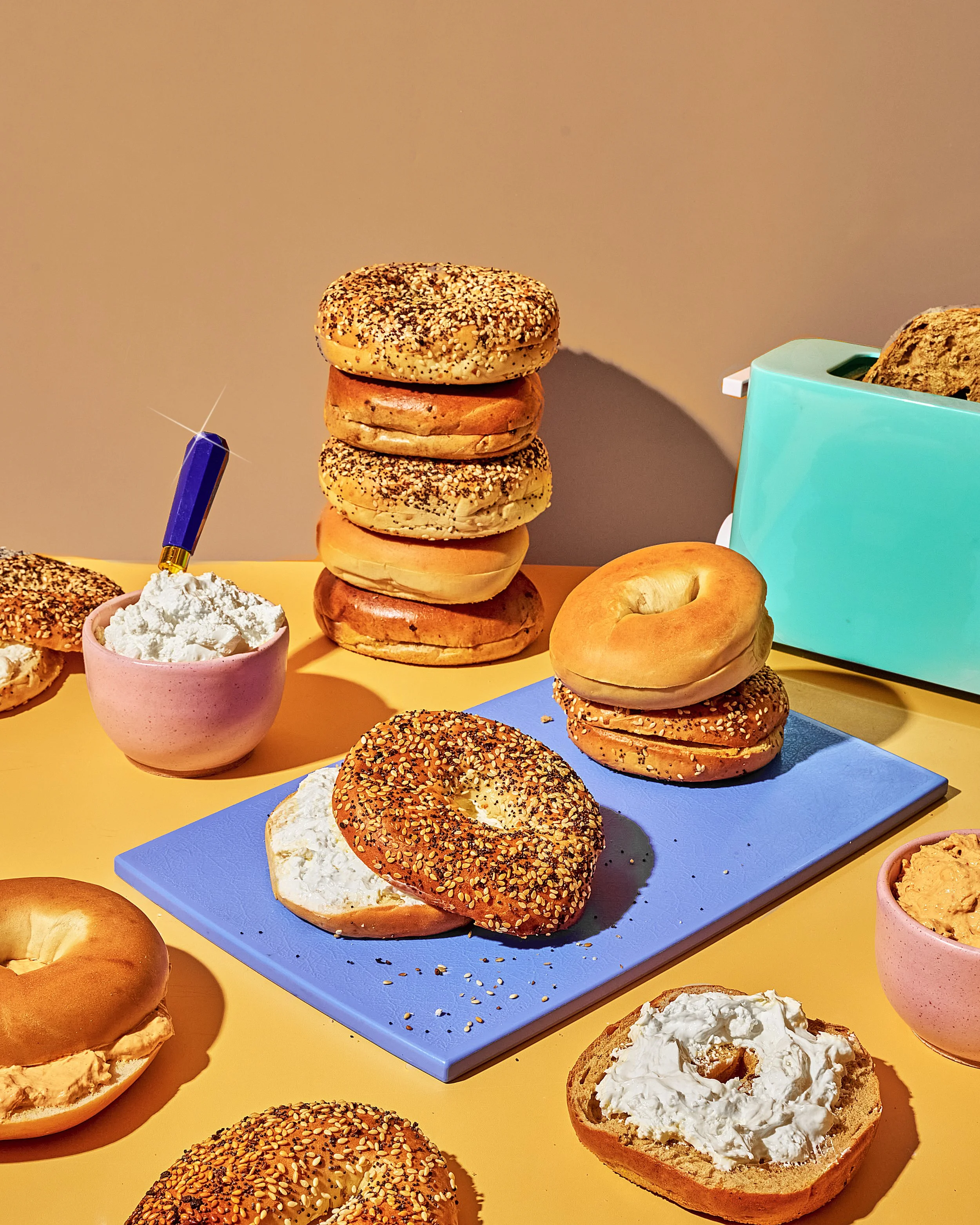 Stacked bagels with sesame and poppy seeds next to a blue cutting board, with open bagel topped with cream cheese in the foreground, and a teal toaster and bowls of cream cheese in the background on a yellow table.