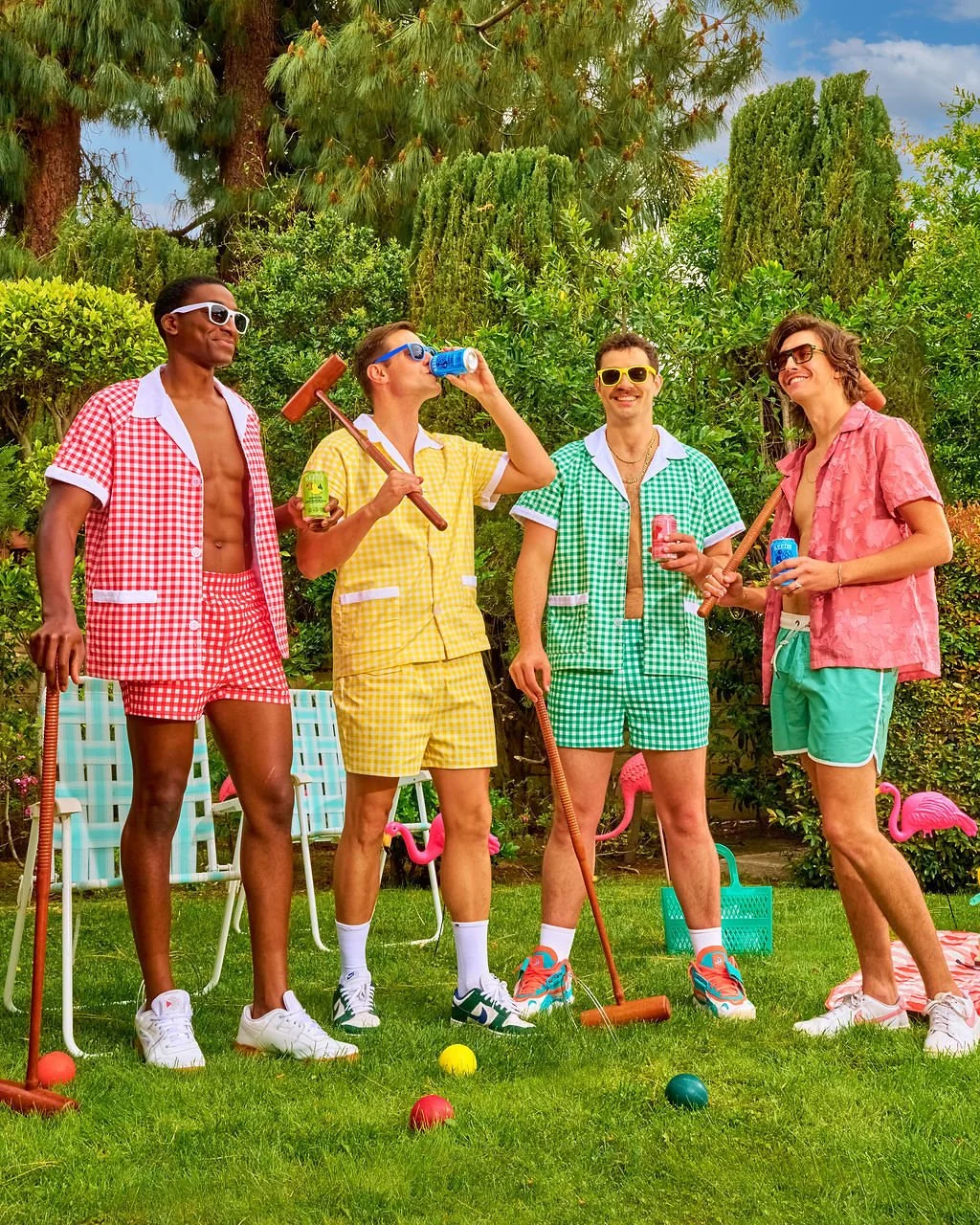 Four young men in colorful shorts and shirts with sunglasses, playing mini-golf outdoors in a lush garden, holding clubs and drinks, with pink flamingo lawn ornaments and a blue sky.