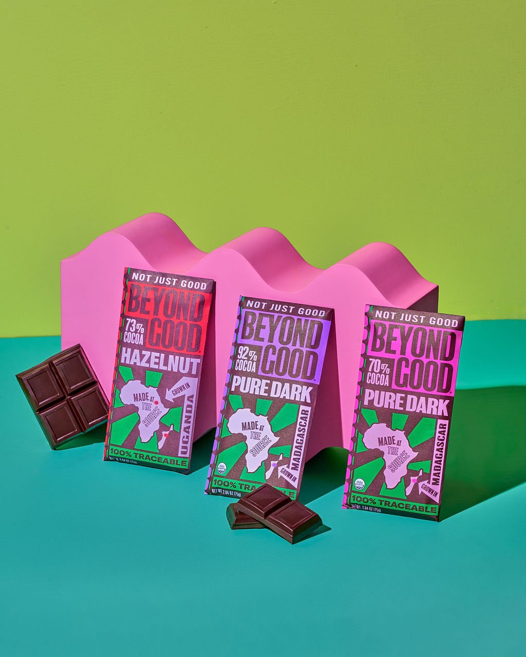 Three chocolate bars with vibrant pink and purple packaging stand in front of a colorful backdrop with undulating pink shapes. The packaging features a white outline of Africa with the text "Made in the source" and labels indicating different cocoa content and flavors, such as hazelnut, pure dark, and a tropical country. Two chocolate pieces are placed in front of the bars.