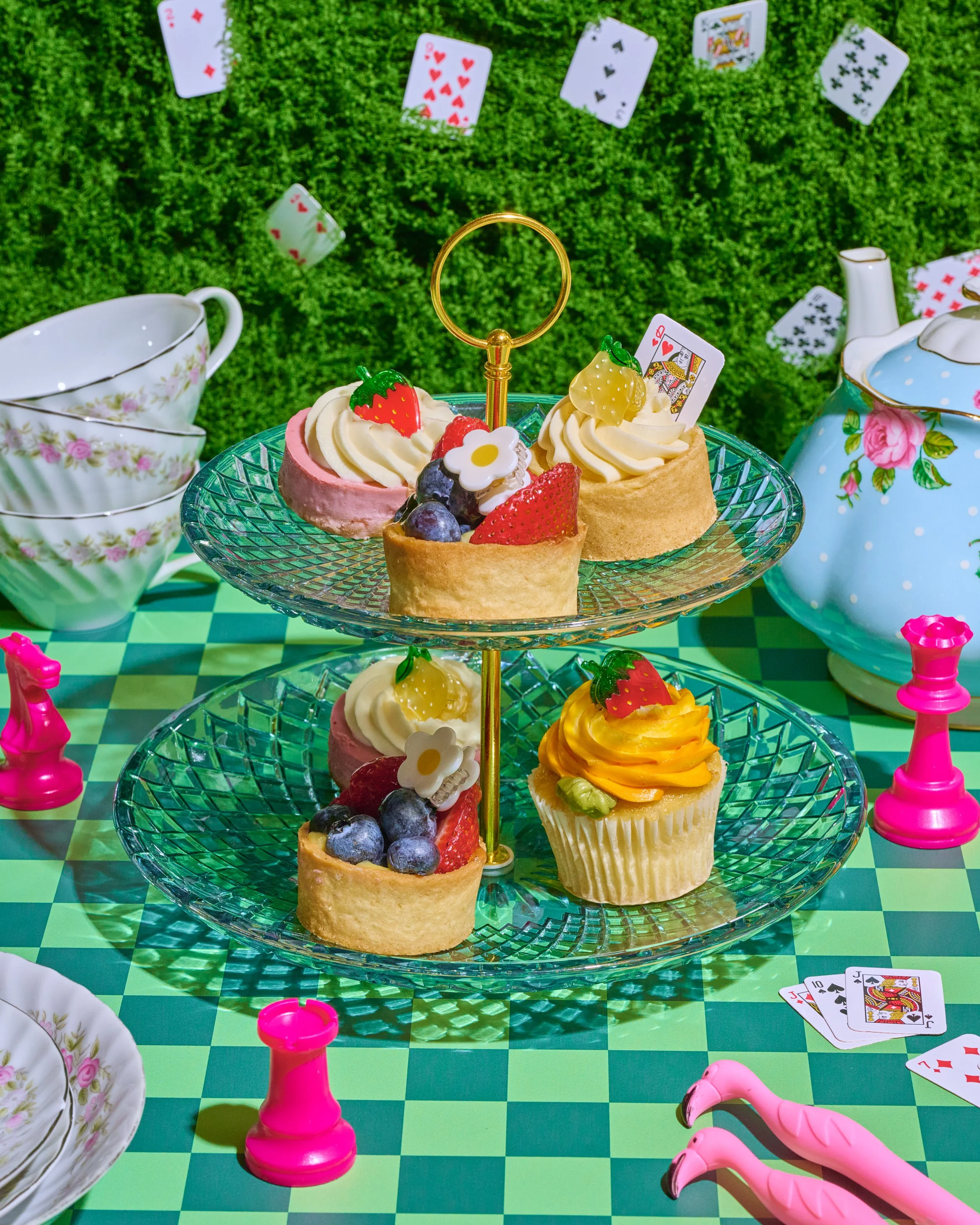 A two-tiered glass dessert stand with assorted fruit-topped tarts and cupcakes, surrounded by playing cards, pink chess pieces, a pink flamingo, and teacups on a green checkered tablecloth, with a green leafy background.