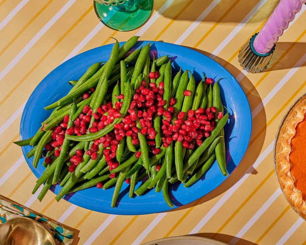 Green beans topped with pomegranate seeds on a blue plate, placed on a yellow striped table.