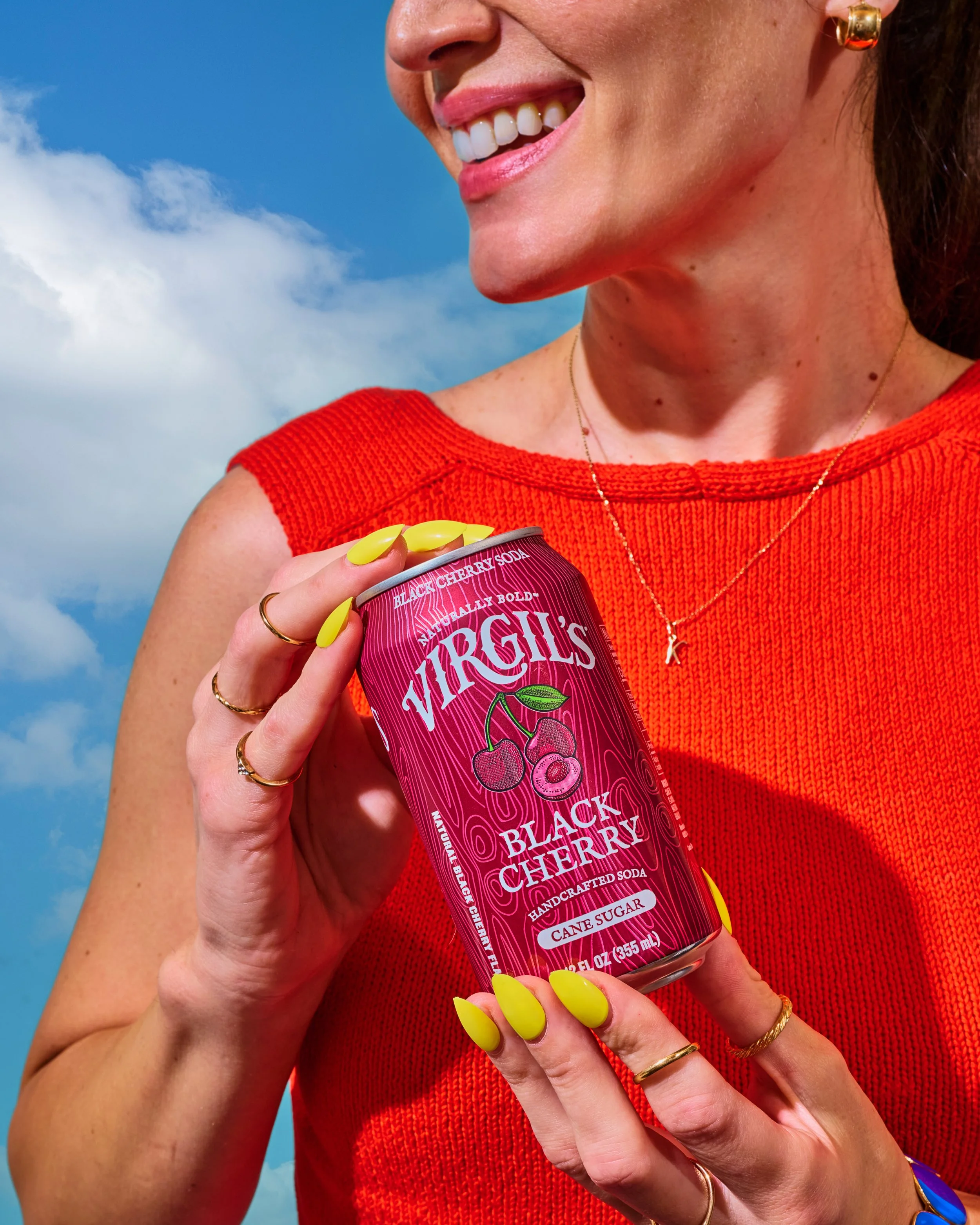 A woman with pink lipstick holding a can of Virgili's Black Cherry soda against a blue sky with clouds. She is wearing a red sleeveless top, gold jewelry, and has bright yellow painted nails.