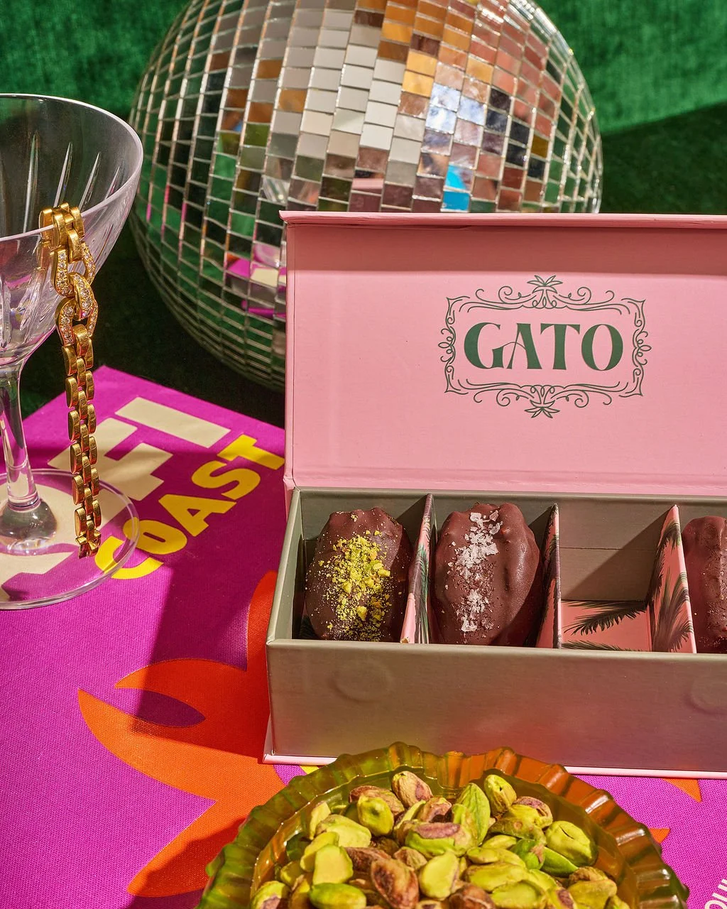 A pink box labeled GATO containing three pieces of chocolate dessert with toppings, a bowl of pistach nuts, a disco ball, a glass with a gold chain, and a tablecloth with colorful text and designs.