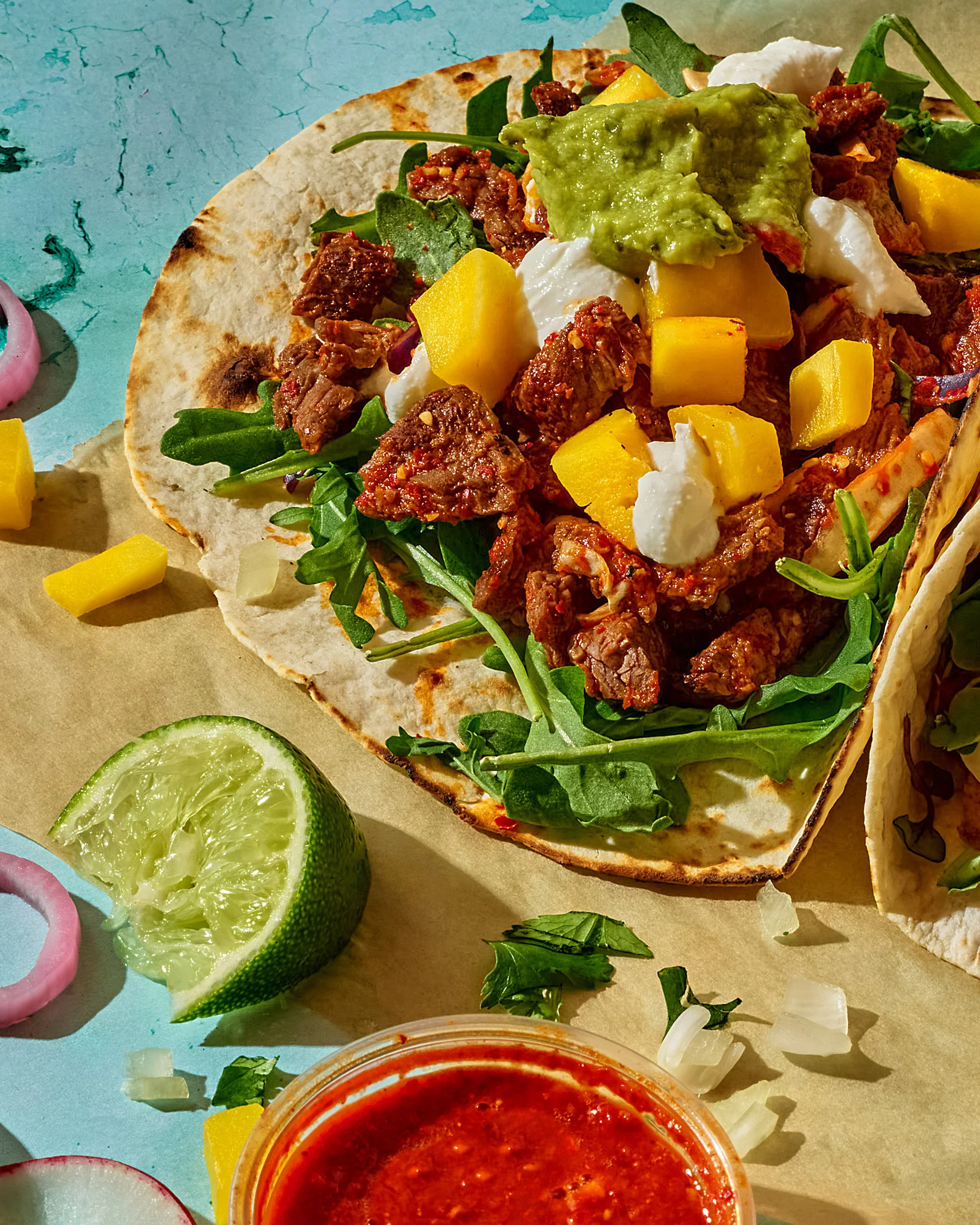 Close-up of a taco filled with beef, cheese, guacamole, and fresh greens, served on parchment paper with lime slices and sides of radish and salsa.