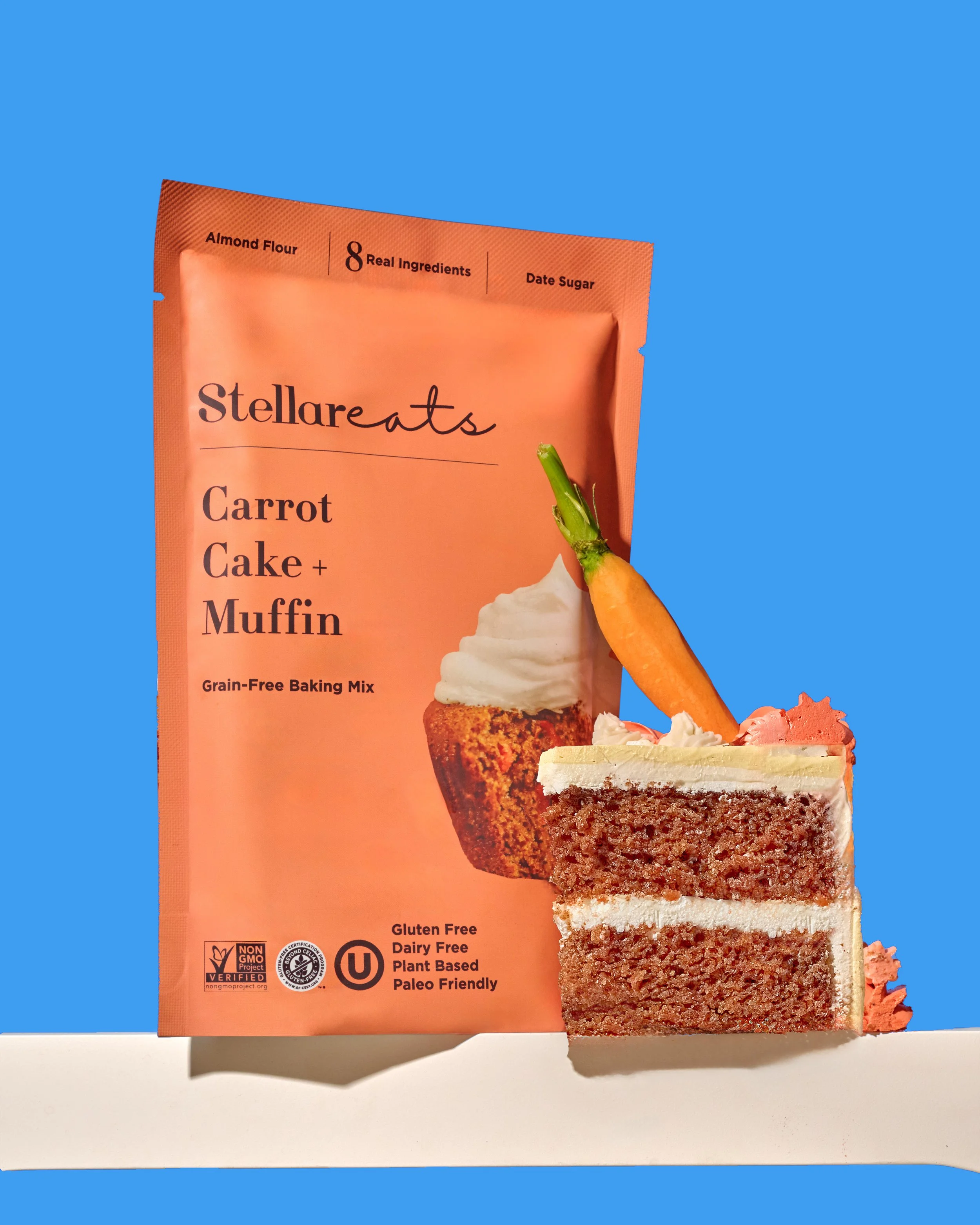 Packet of Stellareats carrot cake muffin mix with a partially sliced piece of carrot cake muffin in front, topped with frosting and a small carrot, against a blue background.