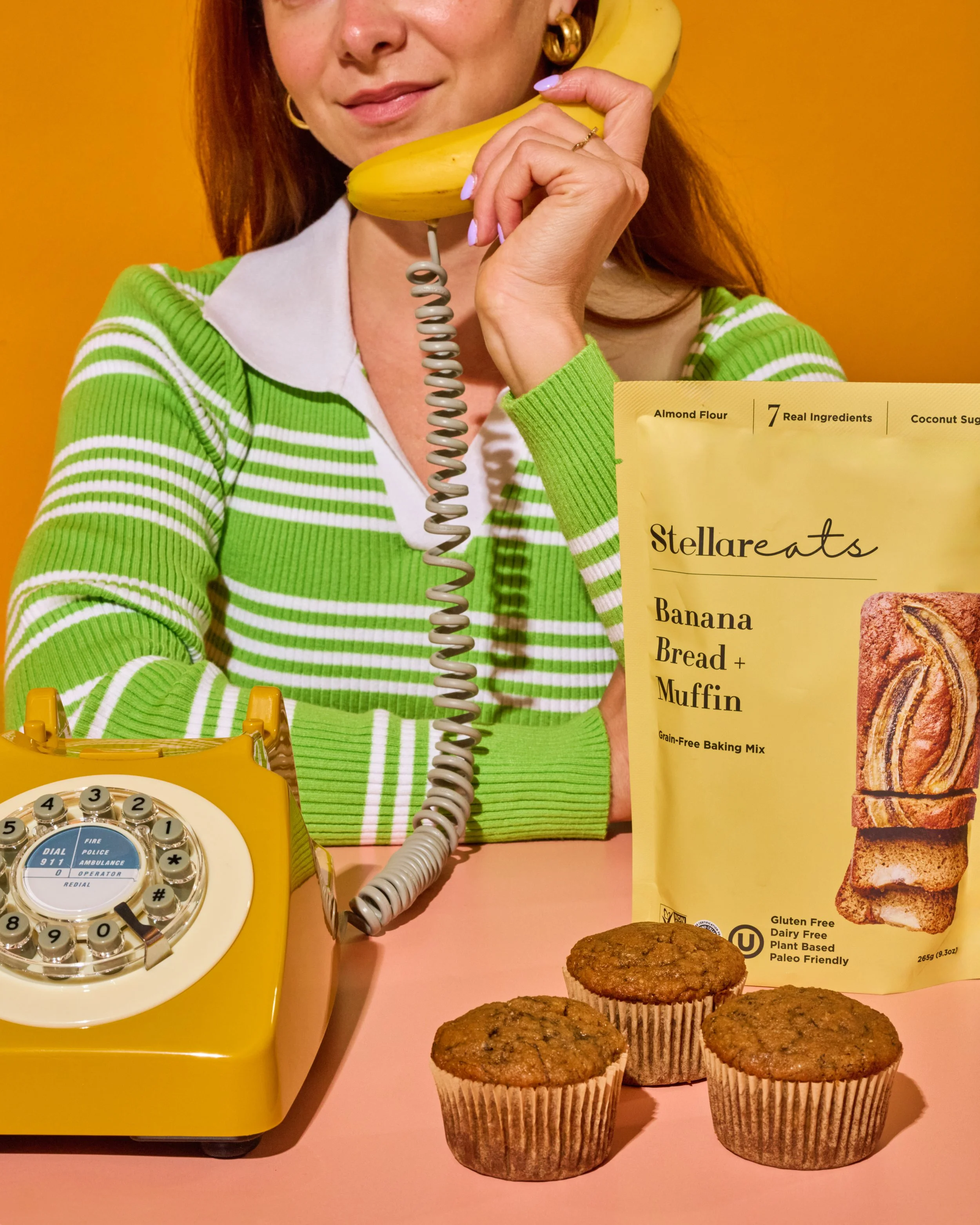 Person with red hair and gold accessories holding a yellow phone to their ear. A yellow rotary dial telephone, a muffin, and a menu featuring banana bread and muffins are on a pink surface against an orange background.