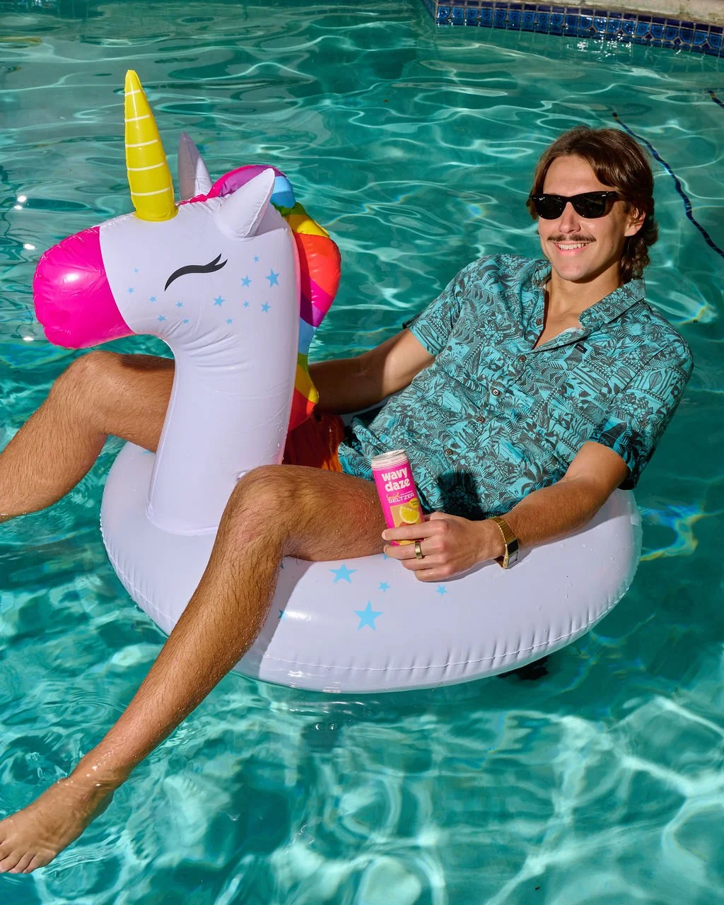 Man relaxing on a unicorn-shaped inflatable in a swimming pool, wearing sunglasses, a patterned shirt, and holding a can of Wavy Daze beverage.