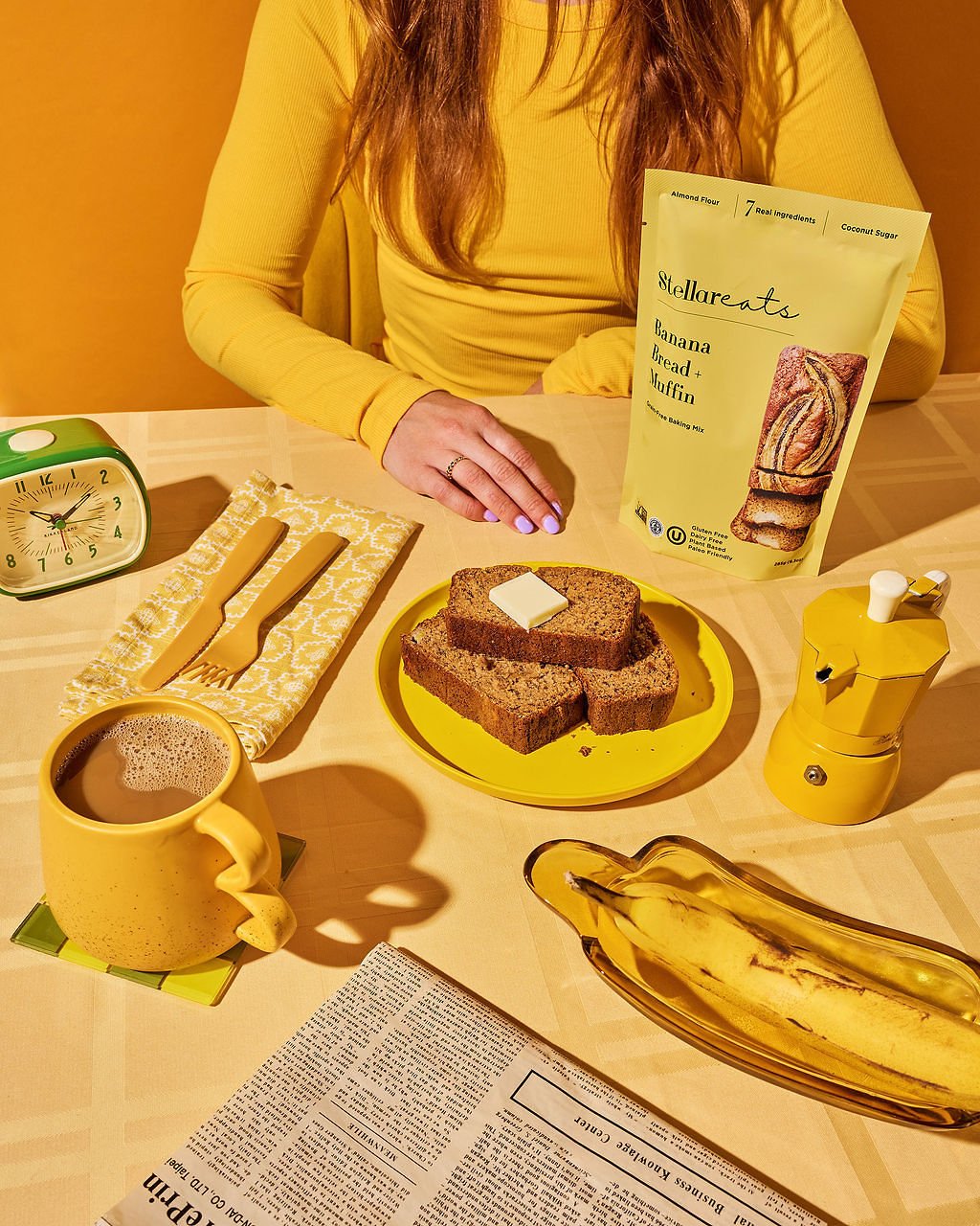A person sitting at a table with a yellow tablecloth, a yellow mug with a hot beverage, a yellow plate with slices of banana bread, a banana on an oval-shaped dish, a spoon and fork on a yellow napkin, a green alarm clock, a yellow coffee grinder, a can of banana bread mix, and a yellow dish with a banana. The person is wearing a yellow long-sleeve shirt.