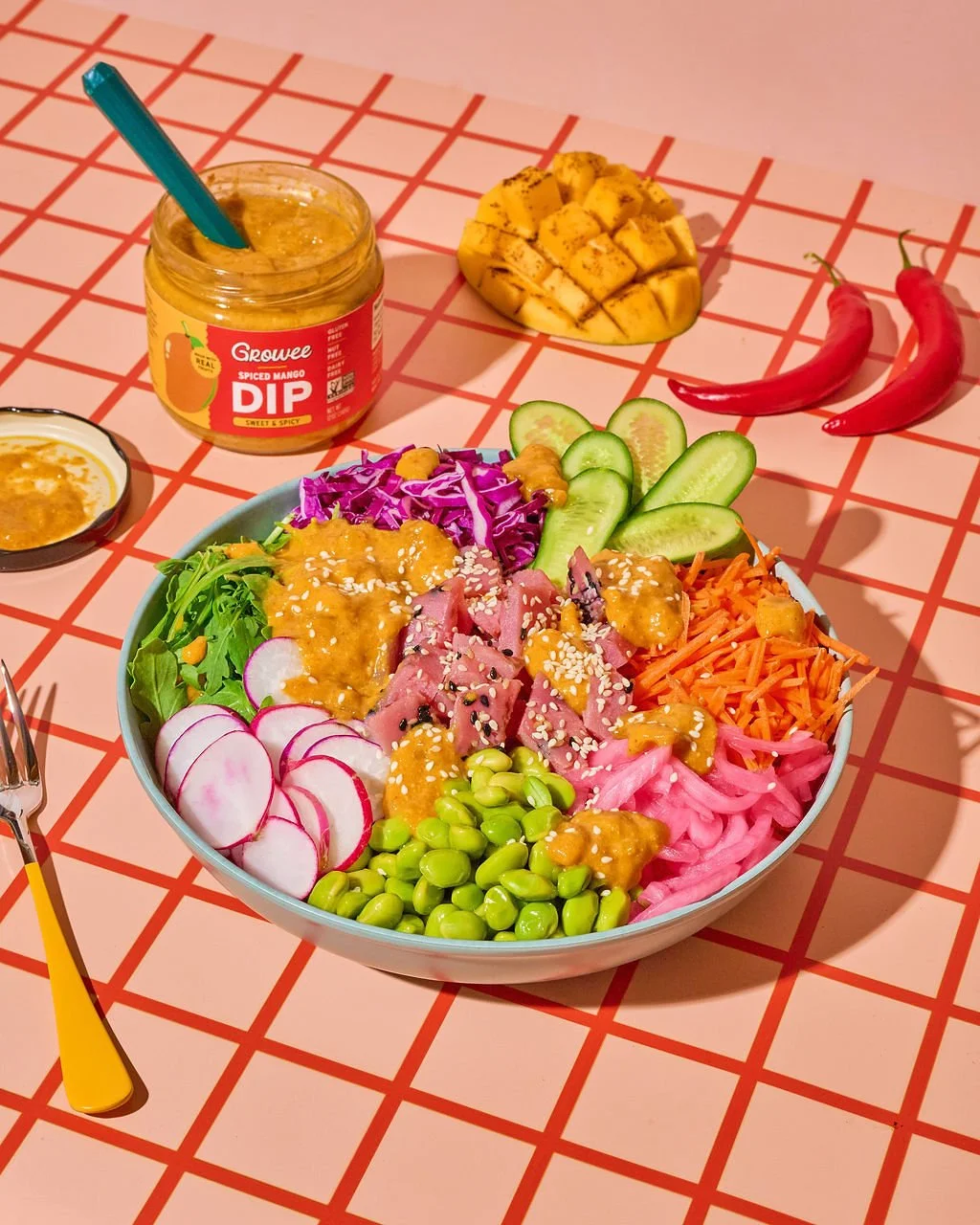 A colorful Asian-inspired salad in a bowl with sliced cucumber, radish, shredded carrots, pickled onions, edamame, purple cabbage, seared tuna, and sesame seeds, garnished with a sauce. Surrounding the bowl are a jar of spiced mango dip, pineapple chunks, two red chili peppers, and a small dish of sauce on a red checkered table.
