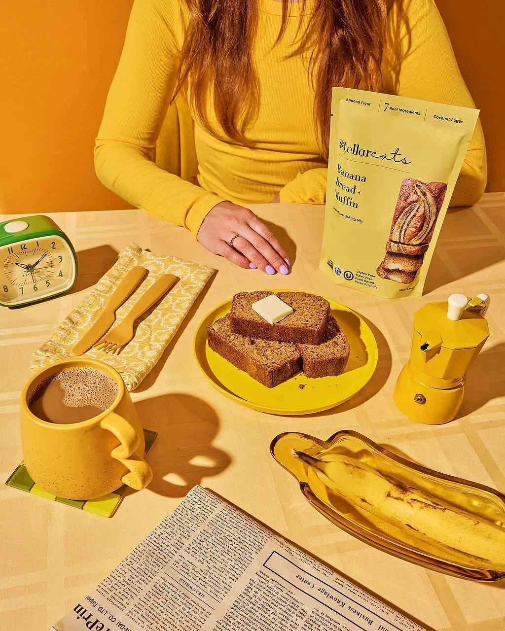 A breakfast scene with a yellow mug of coffee, a banana on a glass dish, a sliced banana bread with a pat of butter on it, and a yellow pepper grinder, on a table with a newspaper, a yellow plate, yellow utensils, and a yellow packet of banana bread 