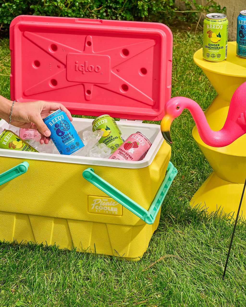 A yellow jug shaped like a flamingo filled with drinks on a green lawn, with a yellow cooler filled with ice and assorted canned beverages, and a pink plastic cooler with a pink lid containing canned drinks.