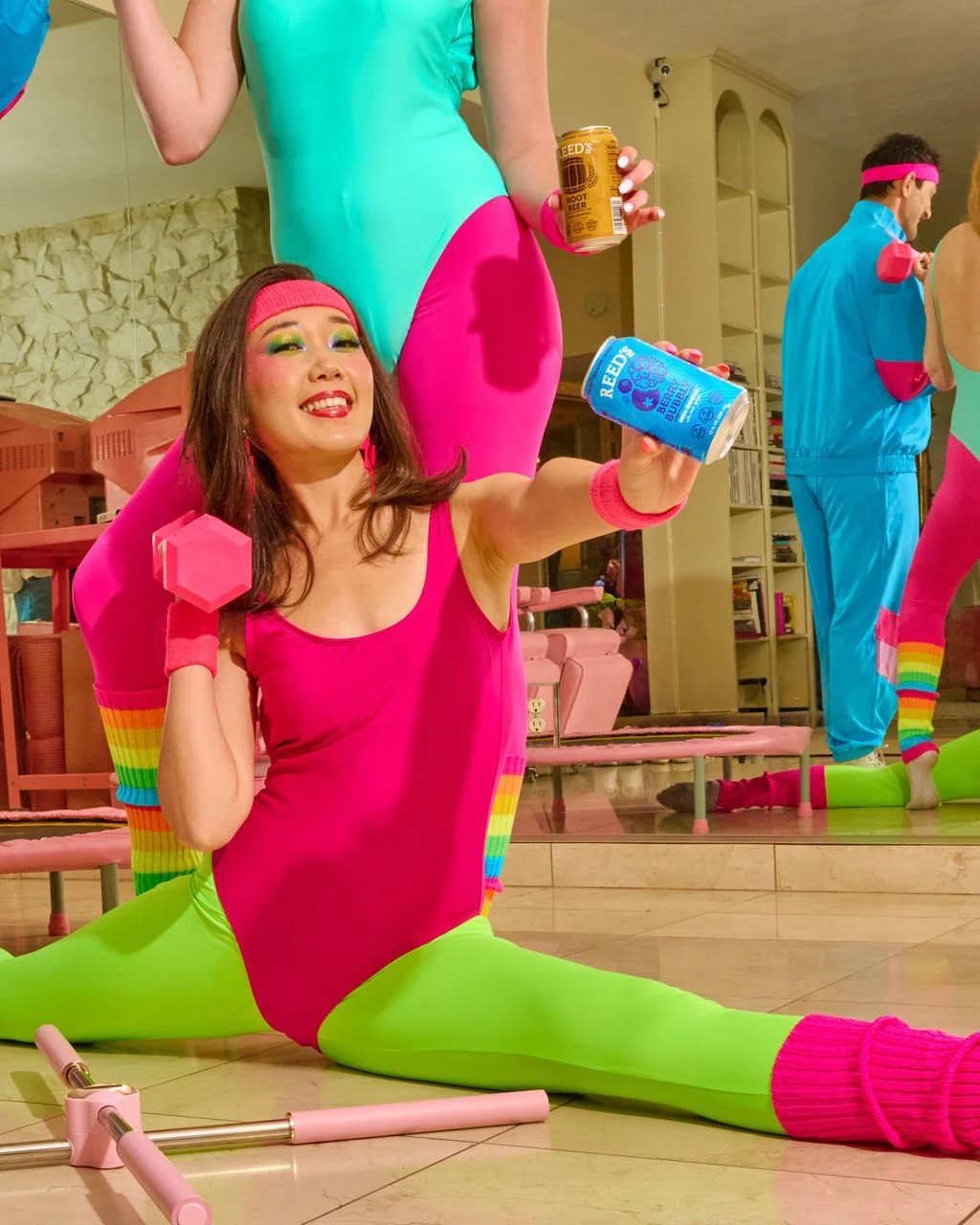 A woman performing a split yoga pose in colorful workout attire, holding a can of Reed's Ginger Beer, with a brightly decorated gym or studio setting in the background.