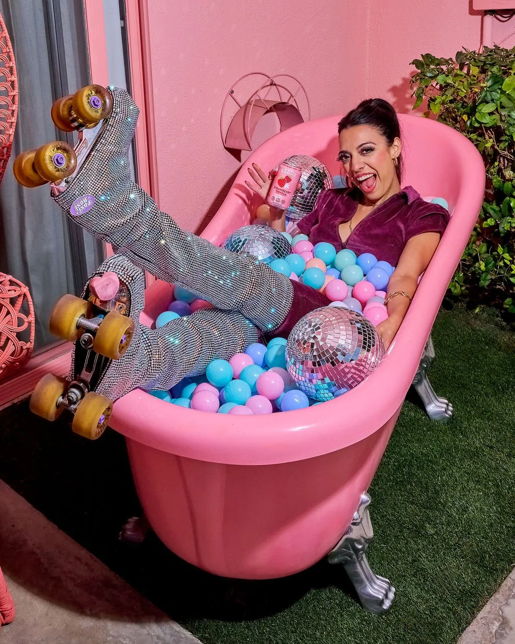 Woman smiling and winking while lying in a pink bathtub filled with pastel-colored plastic balls, disco balls, and wearing sparkly roller skates, holding a drink, with pink walls and decorative items in the background.