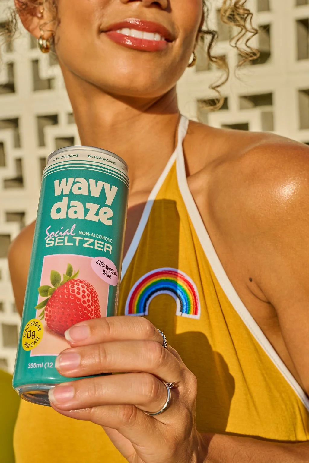 A smiling woman in a yellow top with a rainbow patch, holding a can of Wavy Daze social seltzer with strawberry basil flavor in front of a white patterned wall.