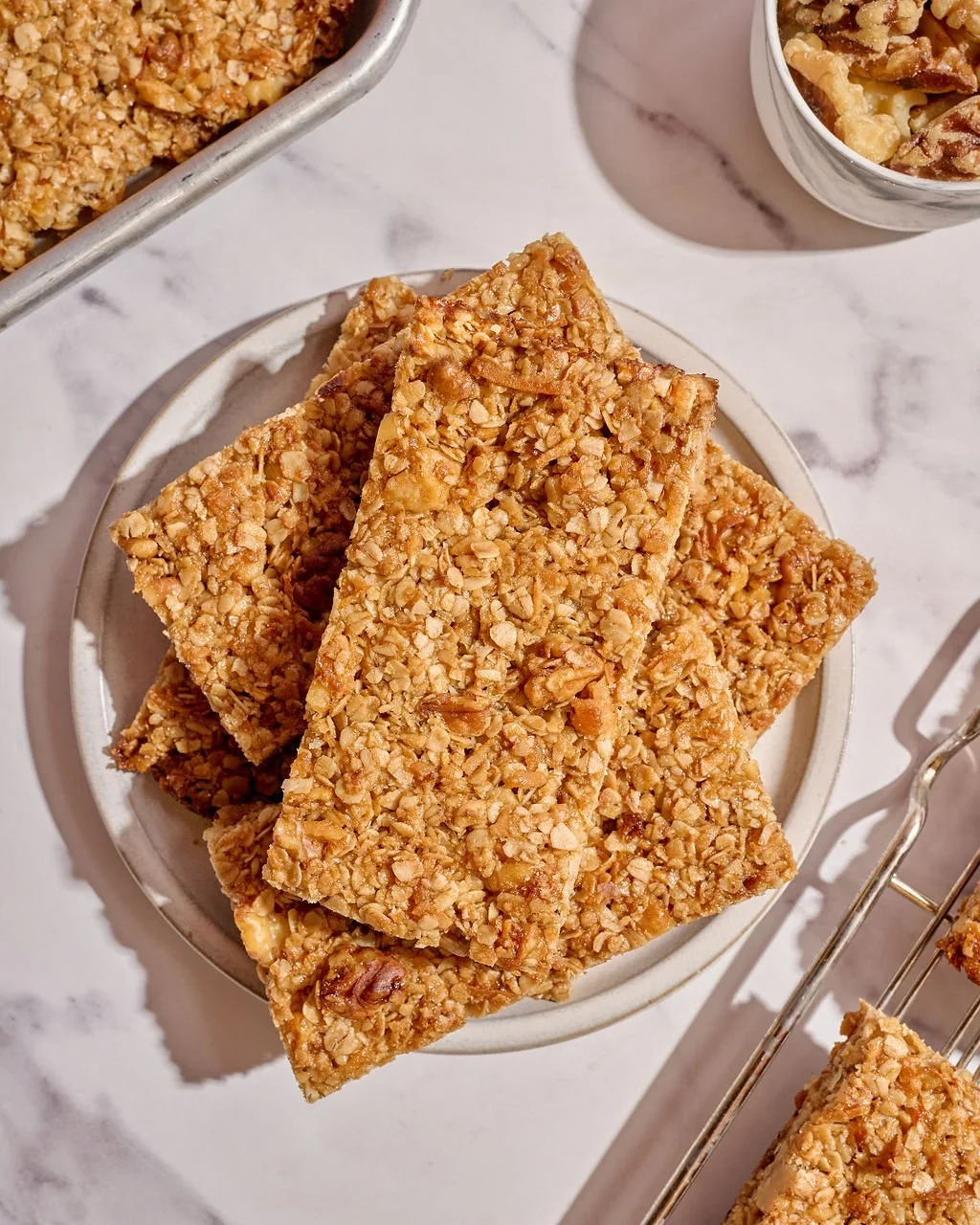 A bowl of granola bars with oats and nuts on a white surface with a glass of granola and a metal tray of more granola bars in the background.