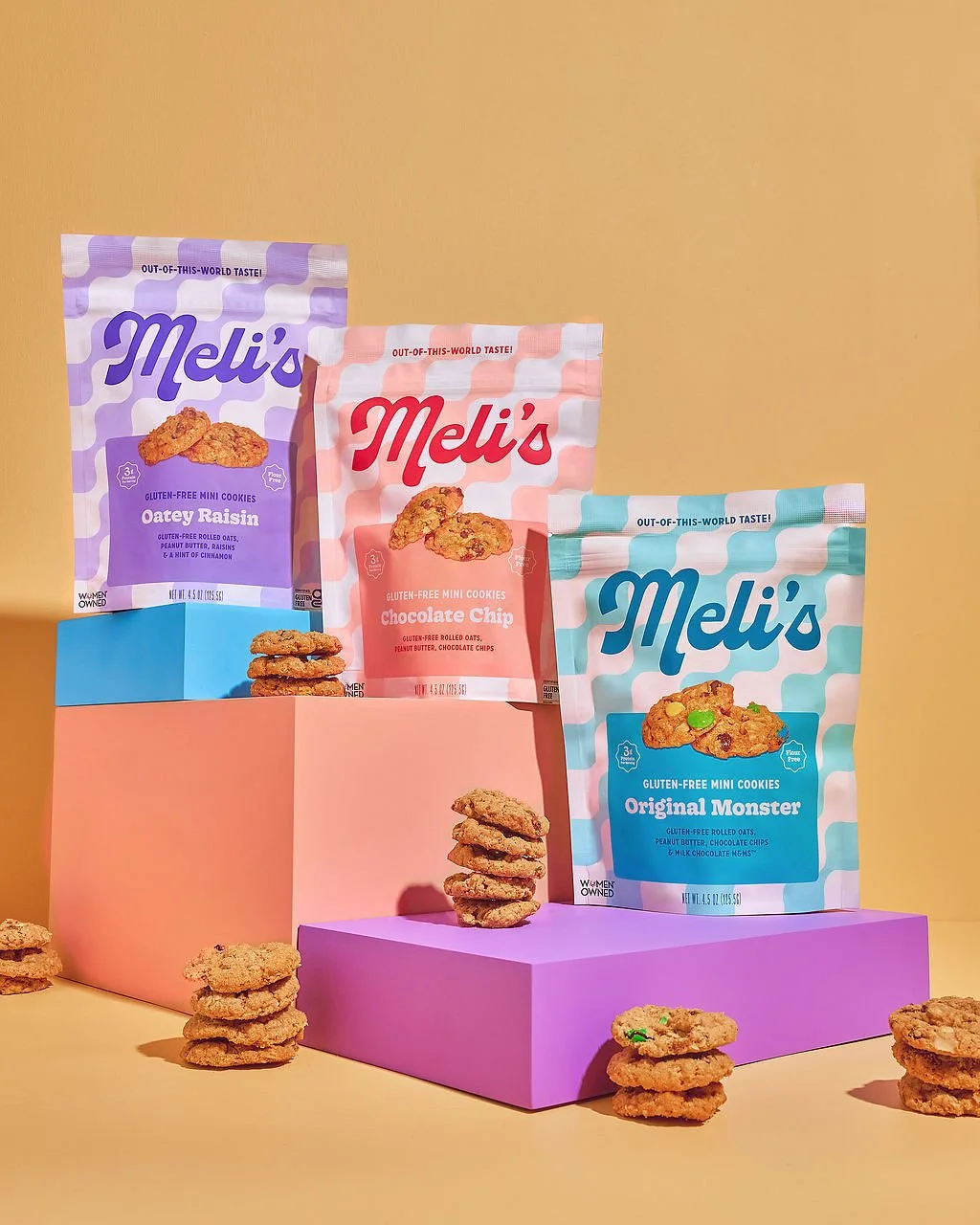 Three bags of Meli's gluten-free mini cookies in purple, pink, and blue packaging, with assorted cookies stacked in front of and around the bags, on pastel-colored platforms against a yellow background.