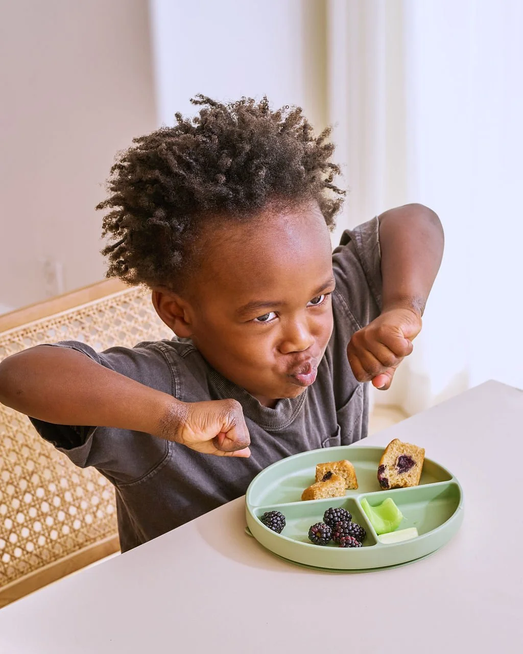A young child with curly hair sitting at a table with a divided plate of snacks, making a playful face and raising fists.