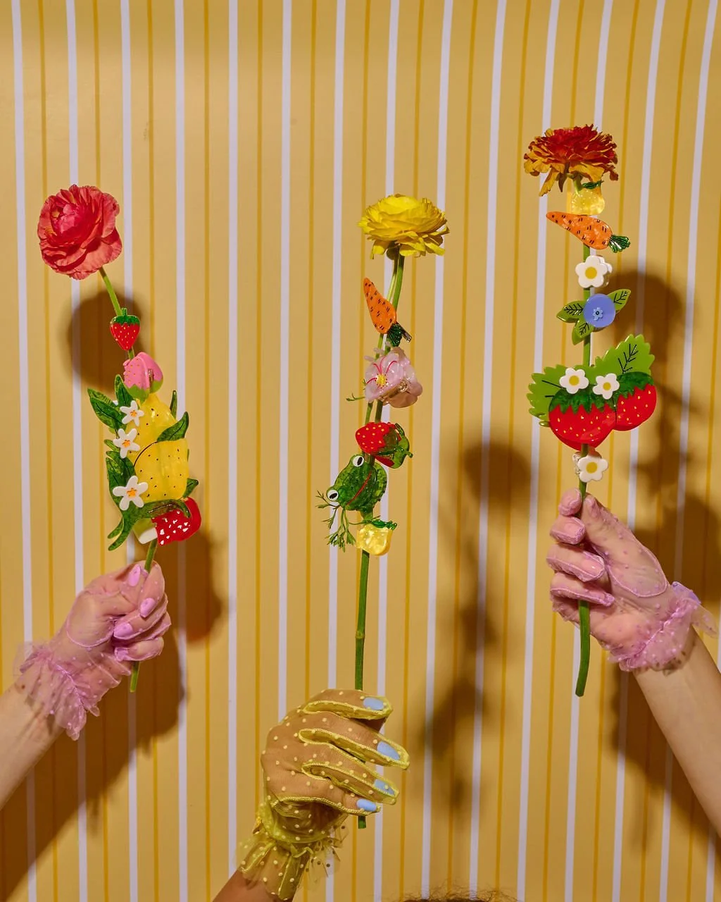 Three hands wearing pink lace gloves holding colorful decorative flower sticks with artificial flowers and small animal and fruit icons in front of a vertically striped yellow and white background.