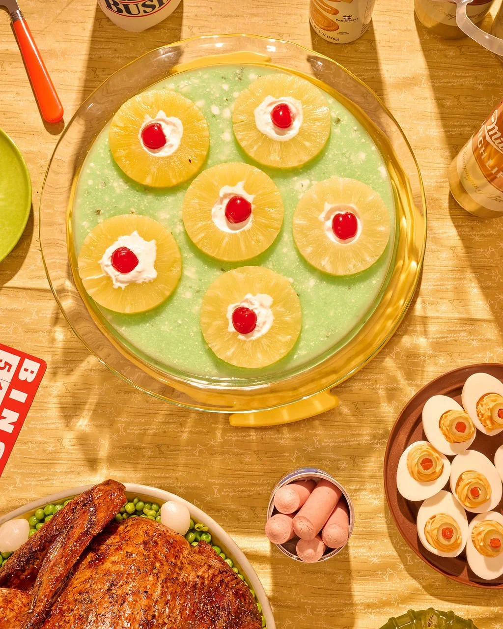 A tray with pineapple upside-down cake topped with maraschino cherries and whipped cream, surrounded by various other dishes including deviled eggs, a roasted chicken with green peas, crackers, and canned beverages on a wooden table.