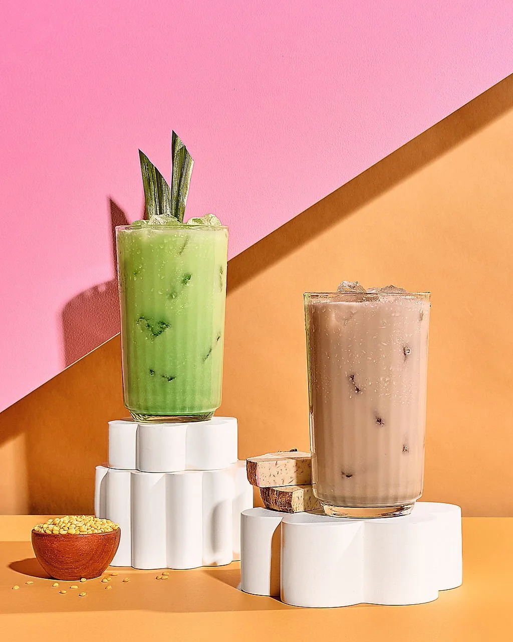 Two vibrant iced beverages in tall glasses on white stackable platforms, with a small wooden bowl of yellow toppings in front, set against a colorful pink and orange diagonal background.