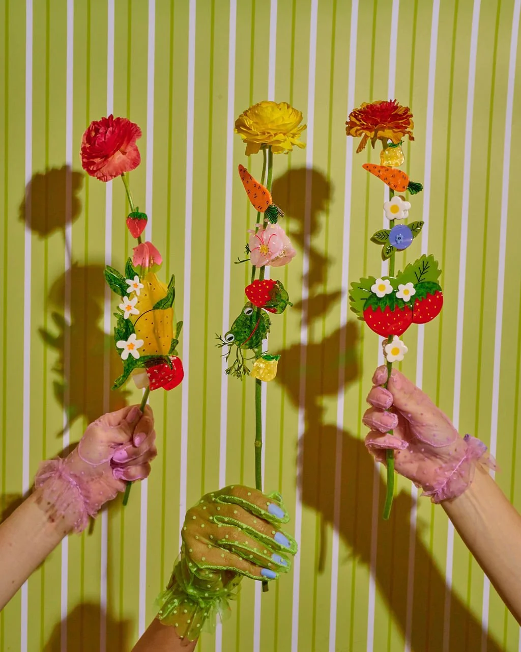 Three people wearing pink gloves are holding colorful toy flower sticks against a green and white striped background.