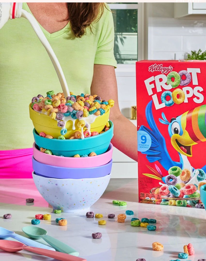 Person pouring milk into a bowl of Froot Loops cereal on a kitchen countertop, with stacked bowls and a box of Froot Loops nearby.