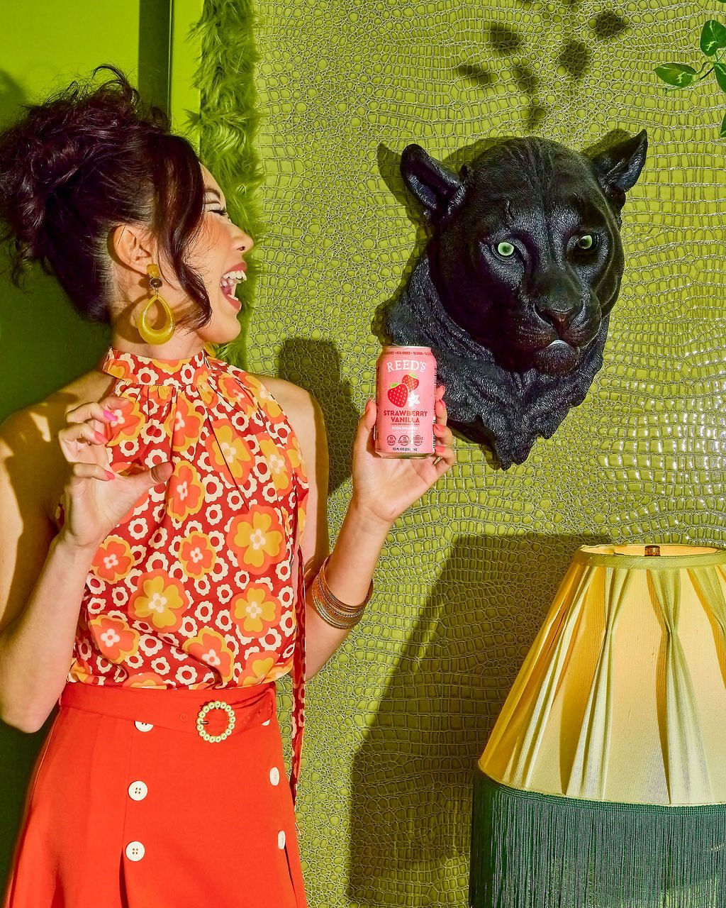 Woman smiling and playfully pretending to hold a can of Reed's Strawberry Vanilla soda next to a black panther head mounted on a green textured wall.