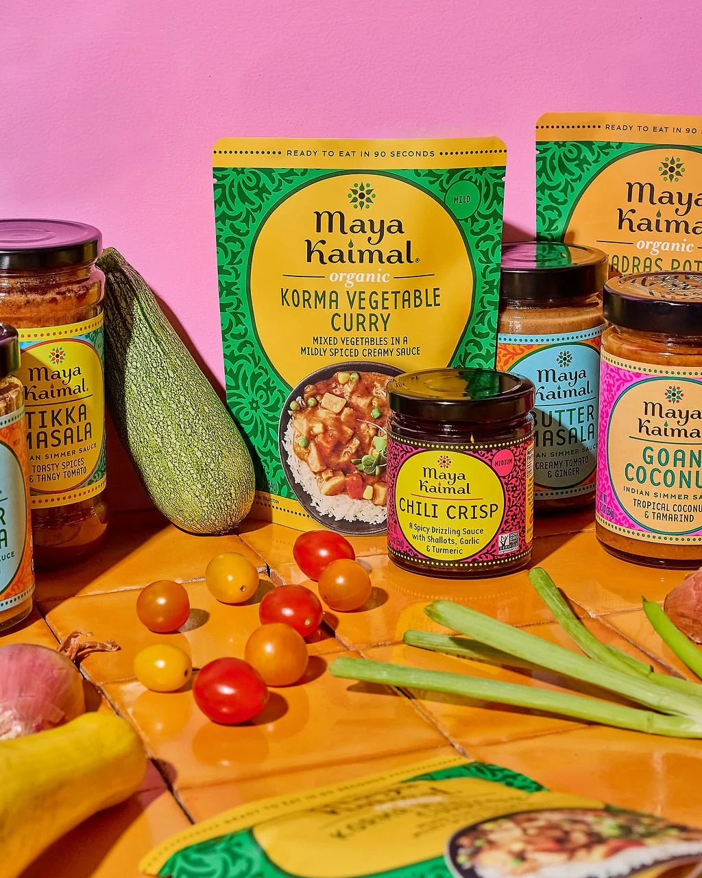 Various jars of Maya Kaimal Indian sauces on a wooden surface, surrounded by vegetables including tomatoes, garlic, green onions, and a zucchini, with a pink wall background.