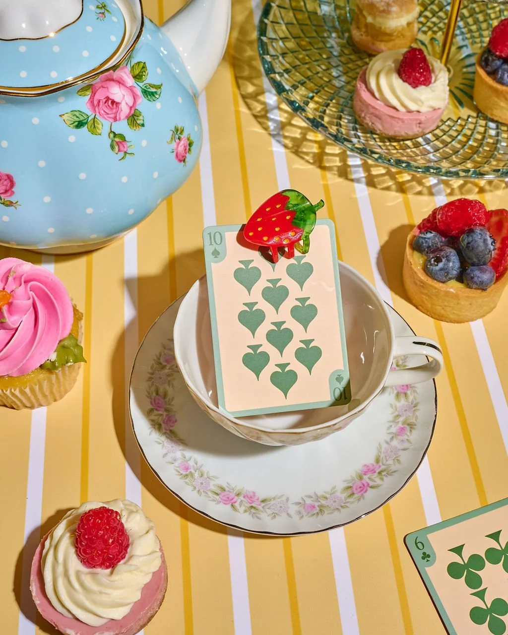 Tea set with cups and saucers, surrounded by small fruit tarts topped with berries, a deck of playing cards, and a teapot, all on a yellow striped tablecloth.