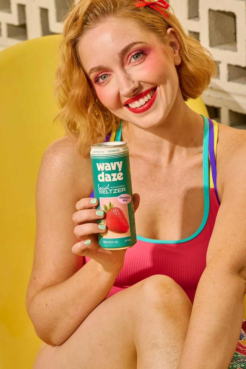 A smiling woman with red hair, pink eyeshadow, and red lipstick holding a can of Wavy Daze Strawberry Basil social seltzer. She is wearing a pink, blue, and purple swimsuit.