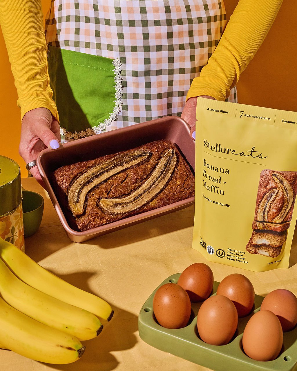 A person wearing a yellow long sleeve shirt and green checkered apron stands behind a wooden table. They hold a baked banana bread in a pink baking dish. On the table are a bunch of bananas, a green egg tray with six eggs, a yellow recipe card titled 'Stellareats Banana Bread + Muffin' with ingredients, and some green bowls.