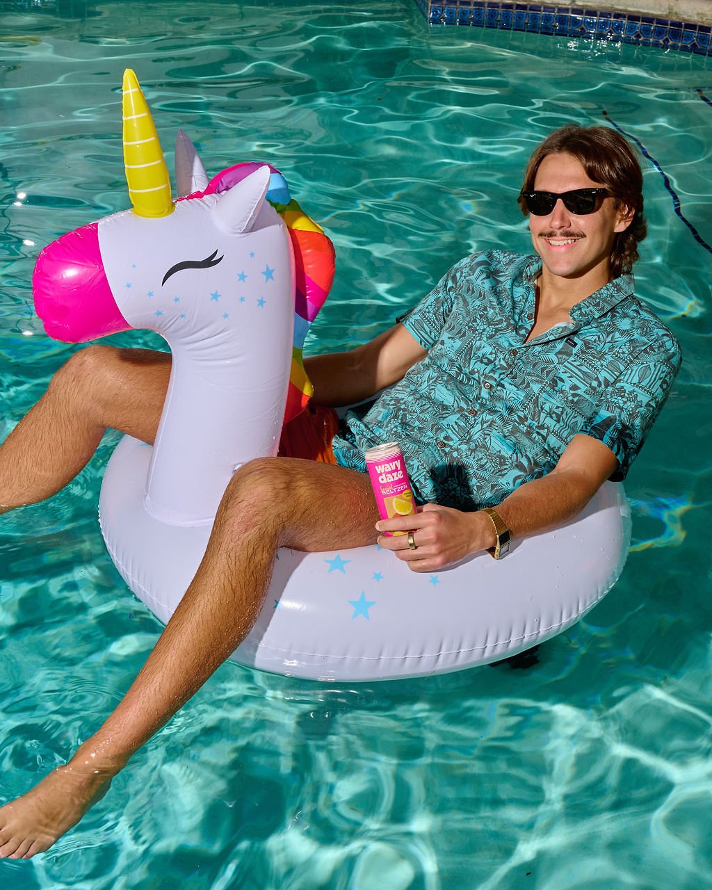 A man with sunglasses and a patterned shirt sitting on a unicorn-shaped inflatable float in a swimming pool, holding a bottle of flavor-boosting spray, smiling at the camera.