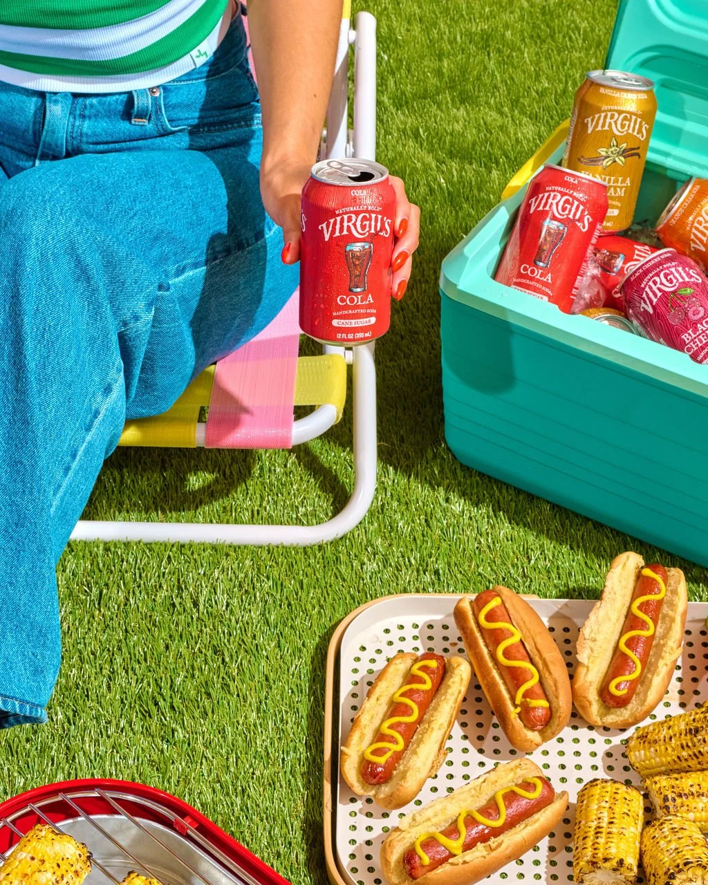Person sitting outdoors on a lawn with hot dogs, grilled corn, and cans of Virgils soda, including cola and vanilla cream flavors.